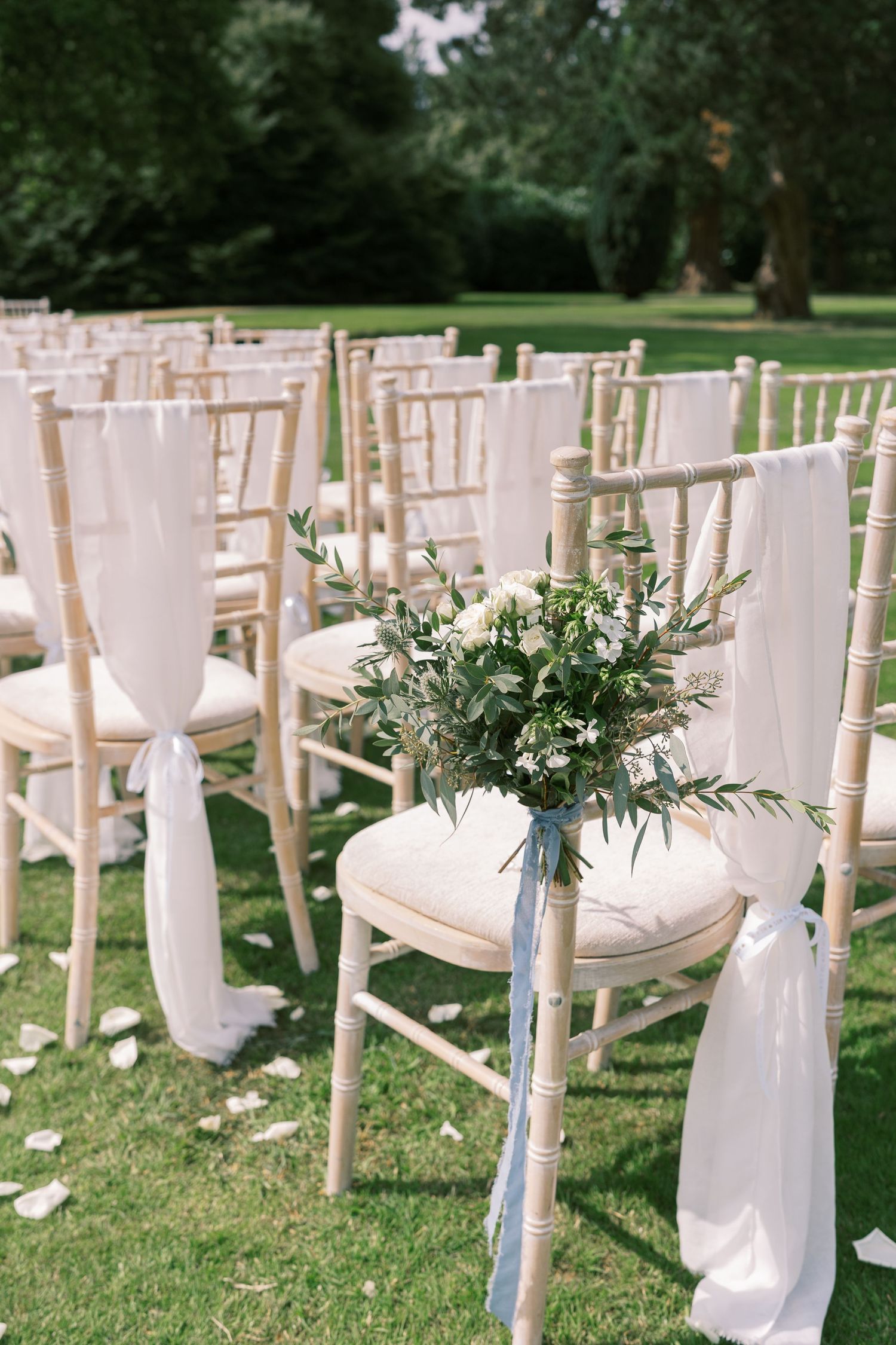 Elegant outdoor wedding ceremony chairs adorned with white fabric sashes and floral bouquets on green lawn.