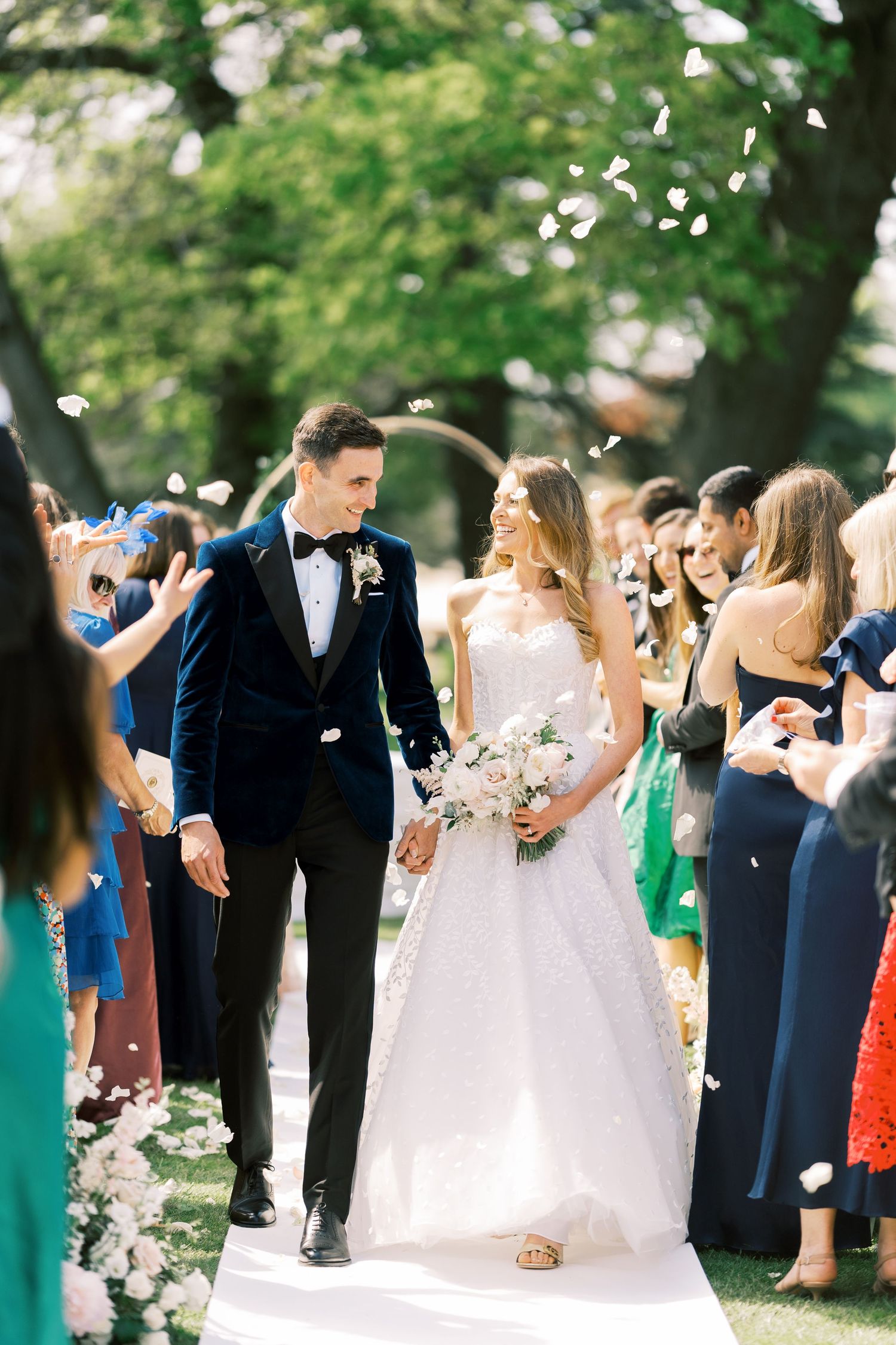 Newlyweds walk down aisle as guests toss white flower petals during sunny outdoor wedding ceremony recessional.
