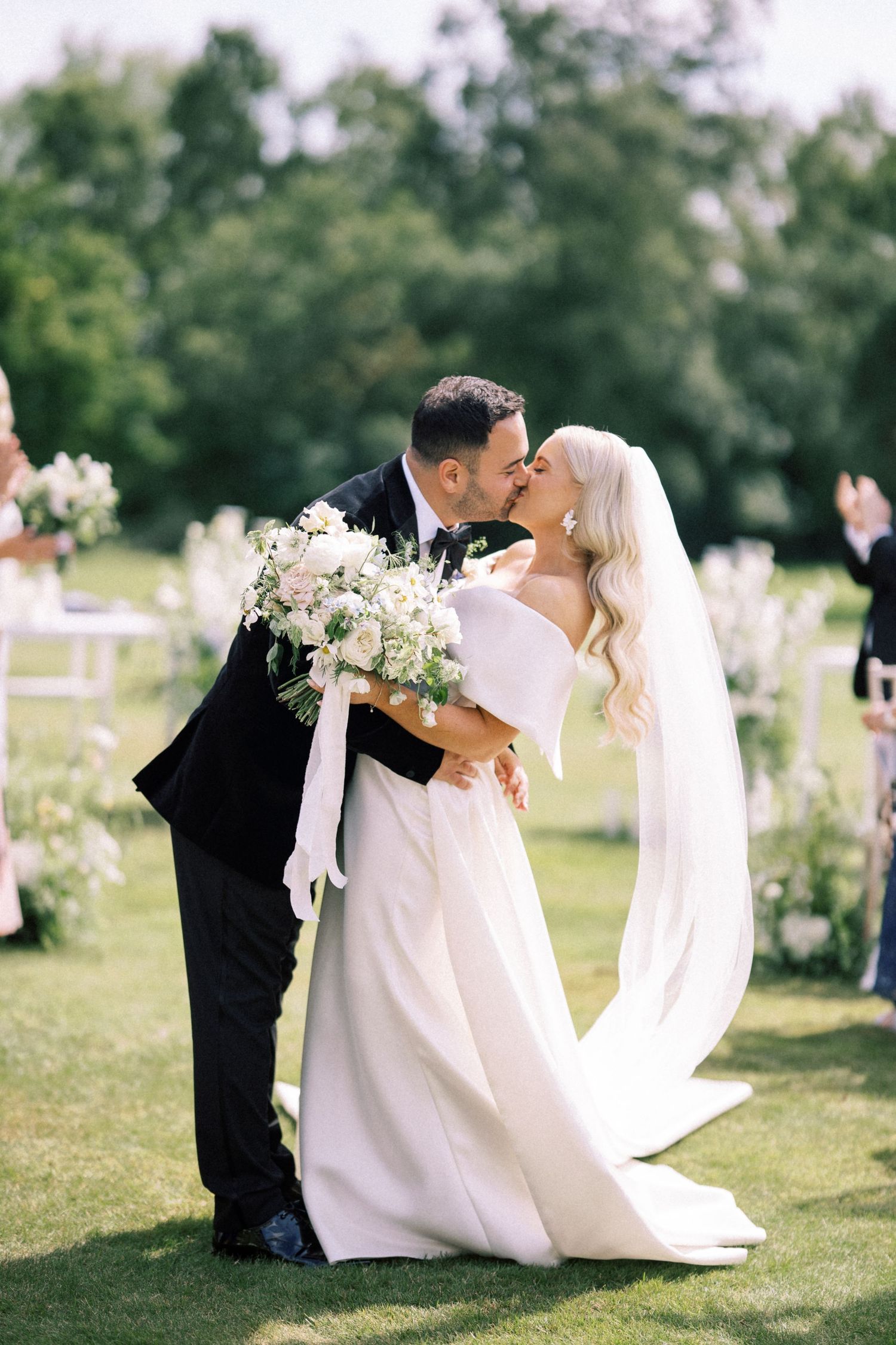 Bride and groom share first kiss outdoors at elegant garden wedding ceremony, bride holding white floral bouquet.