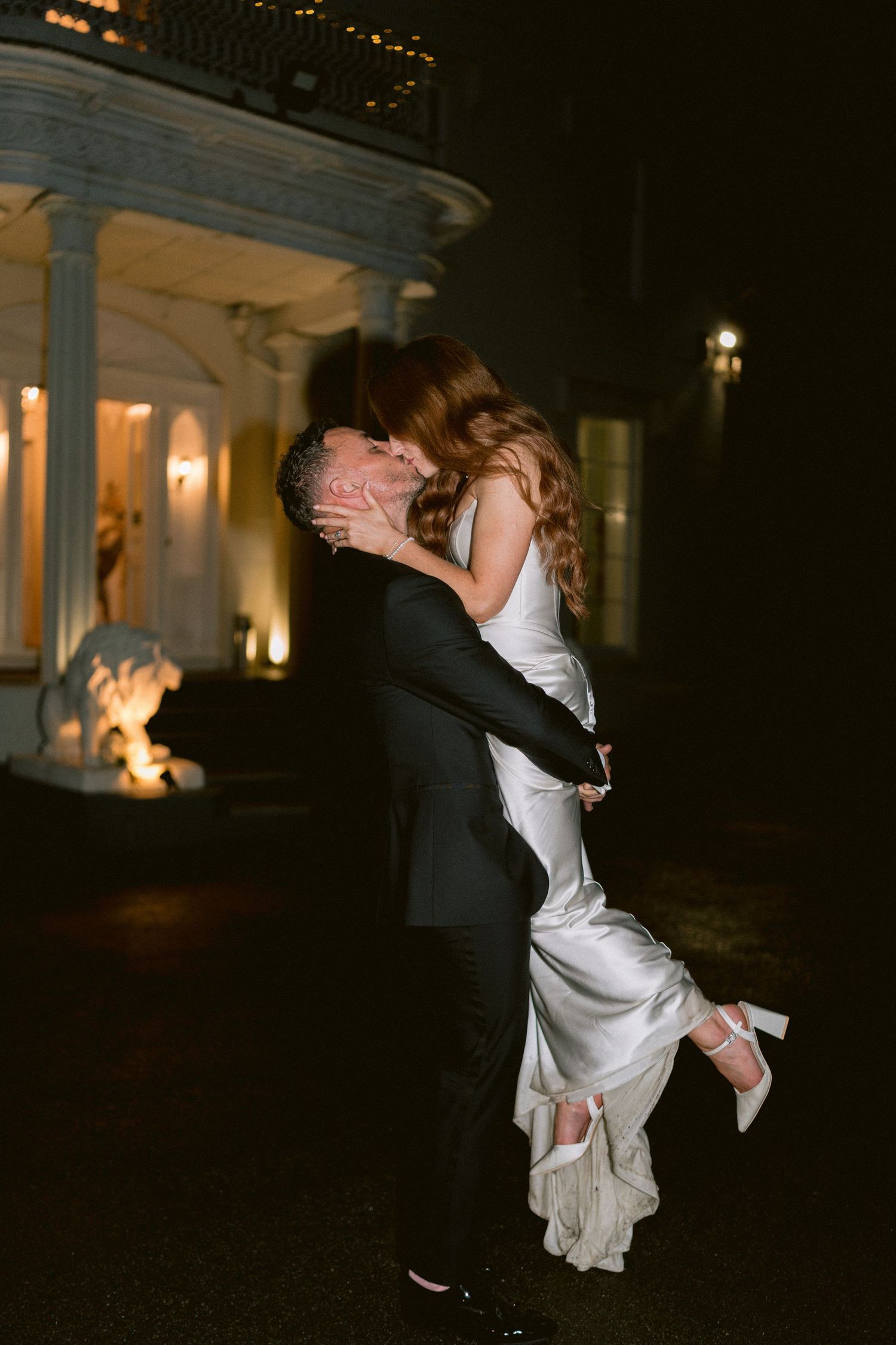 Groom lifts bride in white gown for a romantic kiss outside an elegant lit venue at night.