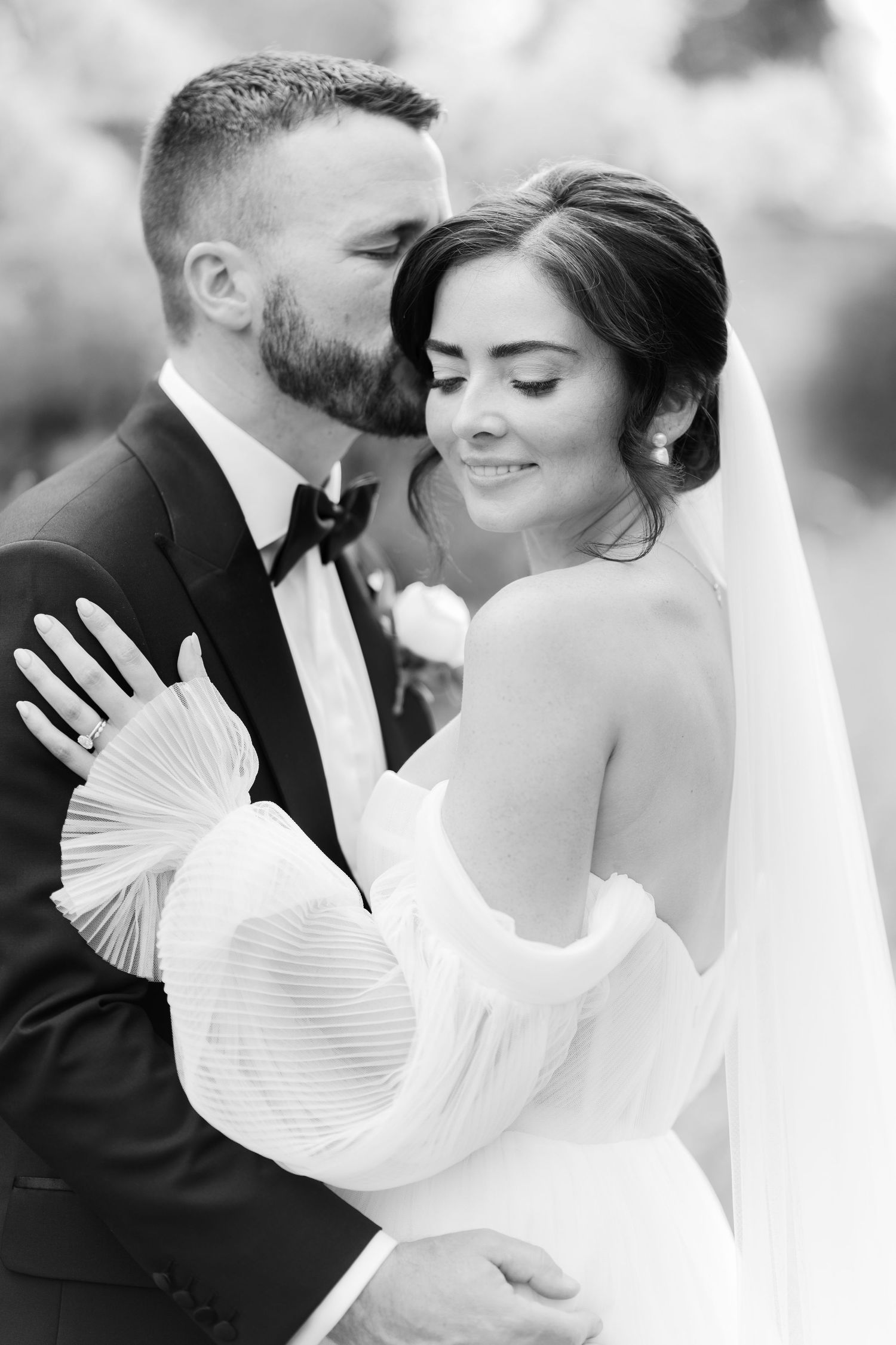 Black and white photo of a groom kissing his bride on the cheek as she smiles in an off-shoulder wedding gown.