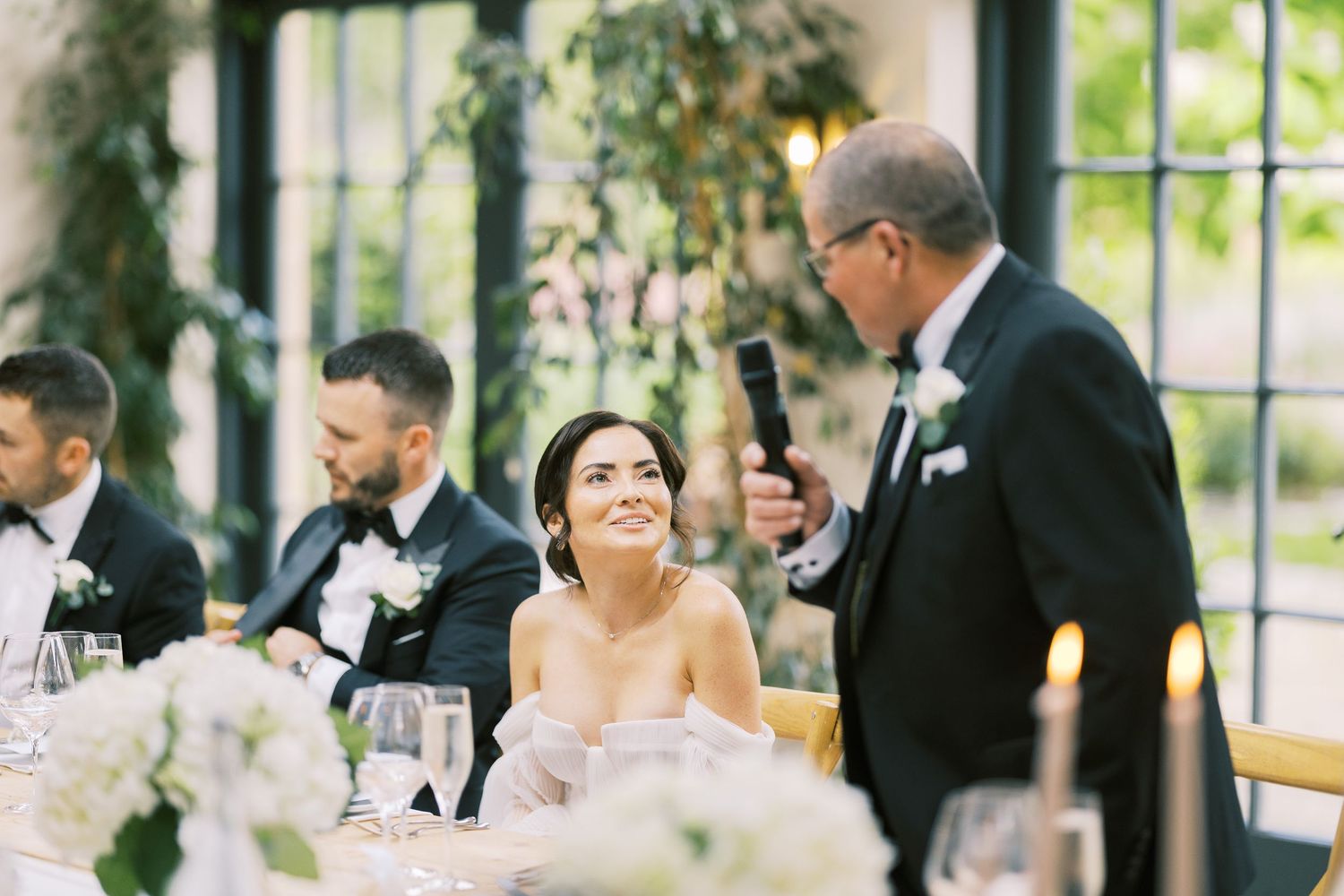Bride smiles warmly as father delivers heartfelt wedding toast at elegant reception with floral decor.