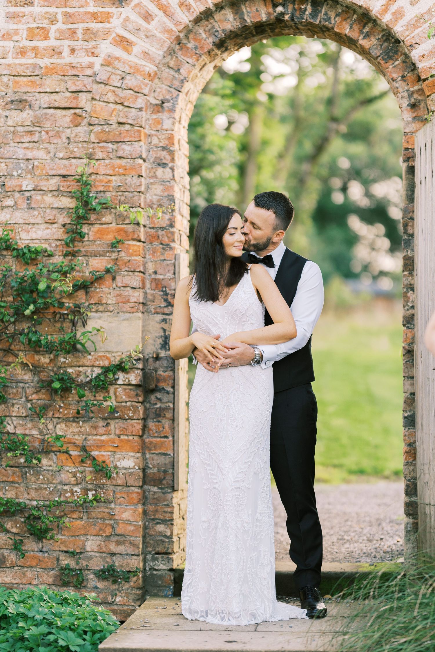 Bride and groom embrace in a rustic brick archway surrounded by lush greenery on their wedding day.