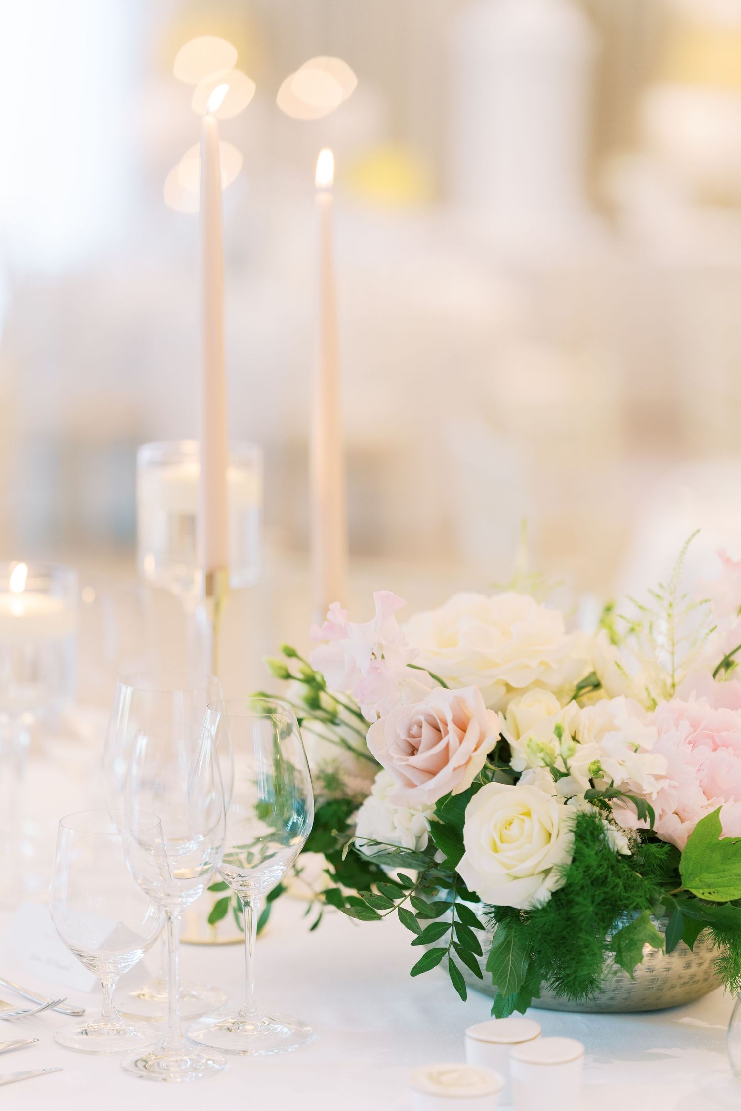 Elegant wedding table centerpiece with blush and white roses, greenery, and tall taper candles on a white linen.