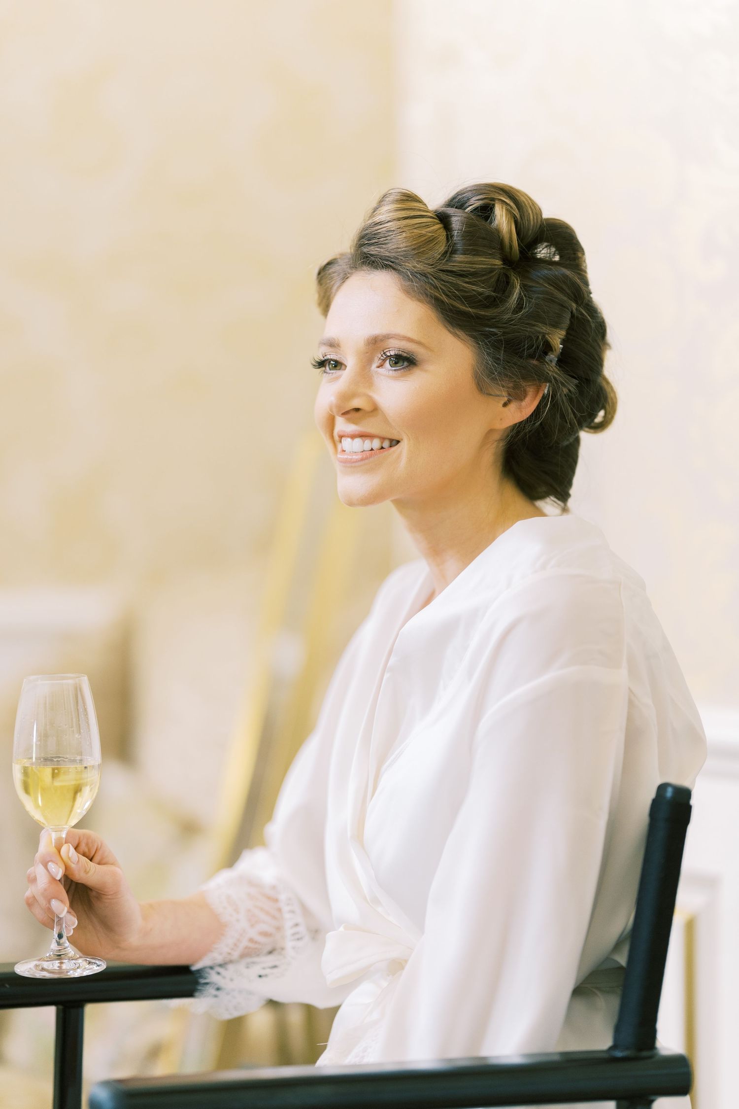 Smiling bride in white robe holding champagne glass with elegant updo hairstyle.