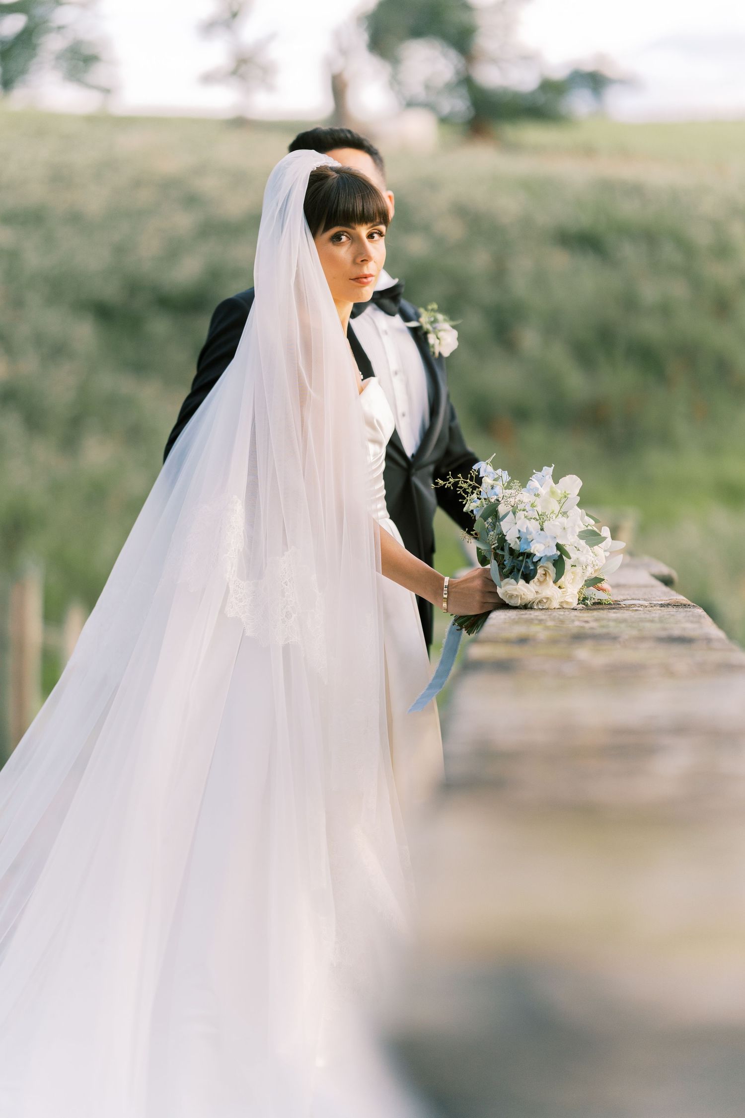 Bride in white gown and veil holds bouquet on rustic bridge, groom in black tuxedo stands behind her.
