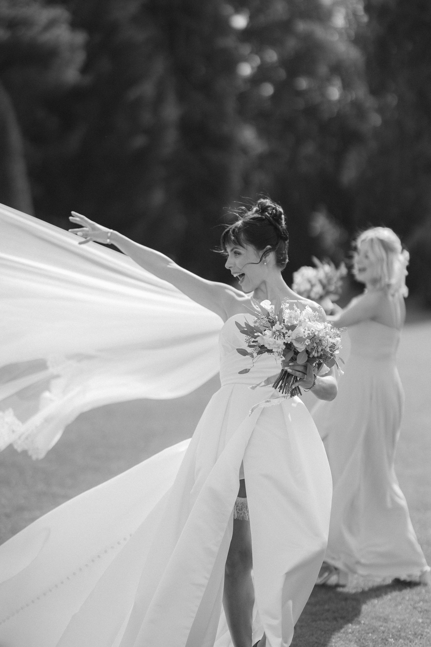Black and white photo of a bride holding bouquet with flowing dress train caught in wind, bridesmaid in background.