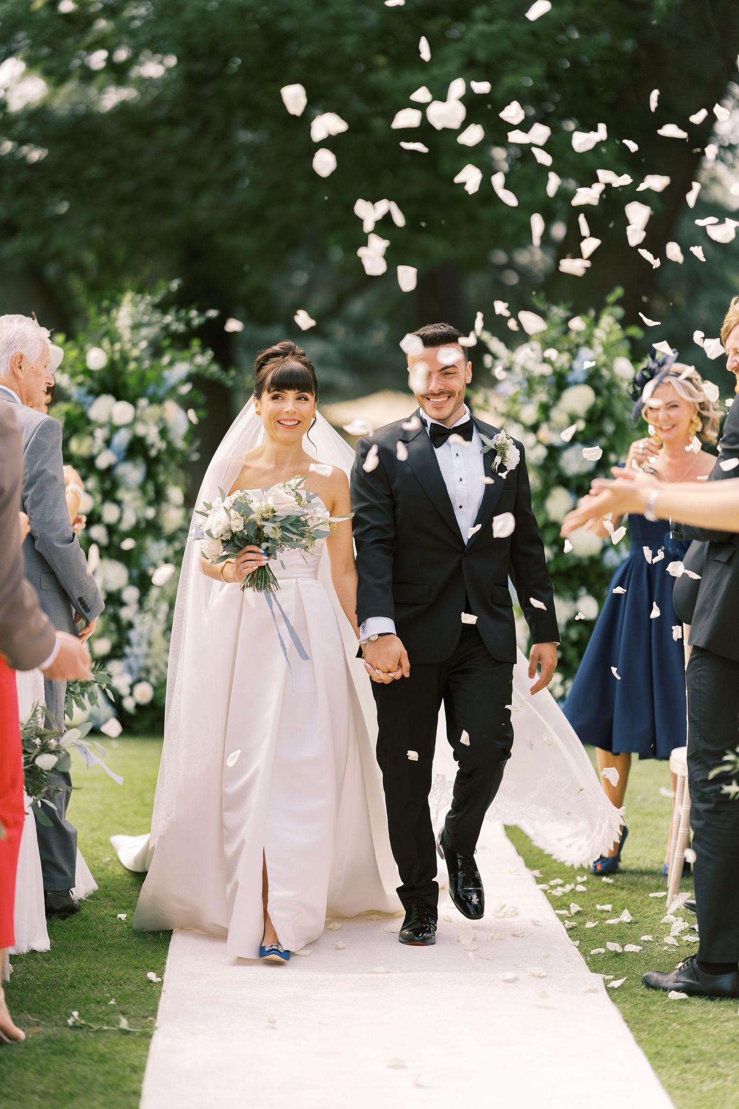 Newlyweds walk down aisle as guests toss white flower petals at elegant outdoor garden wedding ceremony.