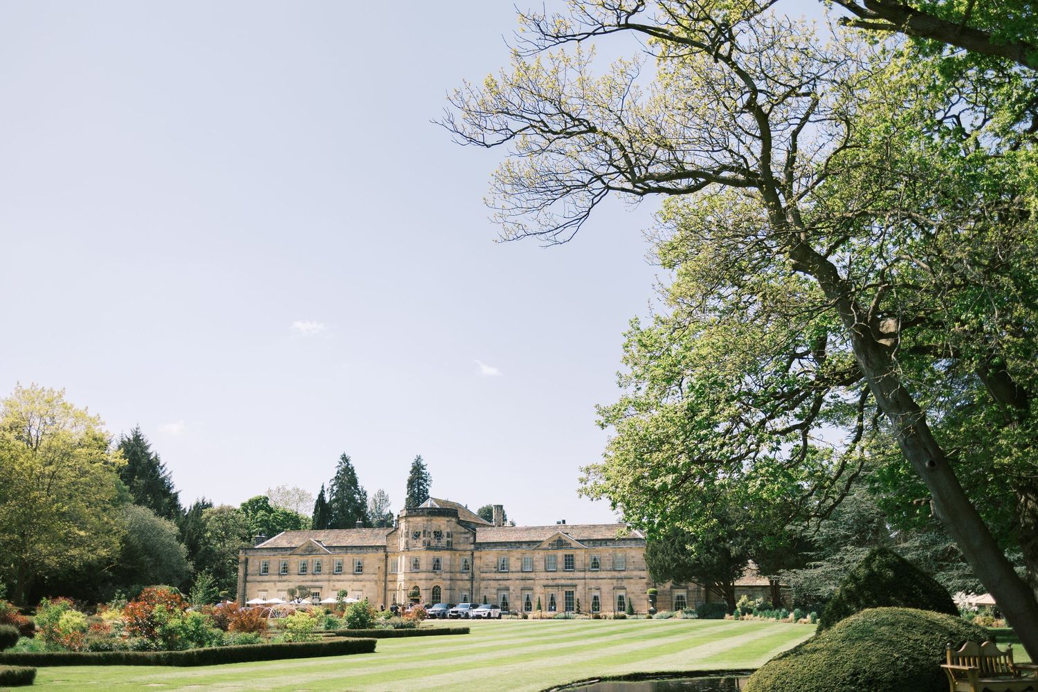 Grand English country manor house with manicured lawns and mature trees under a clear blue sky.