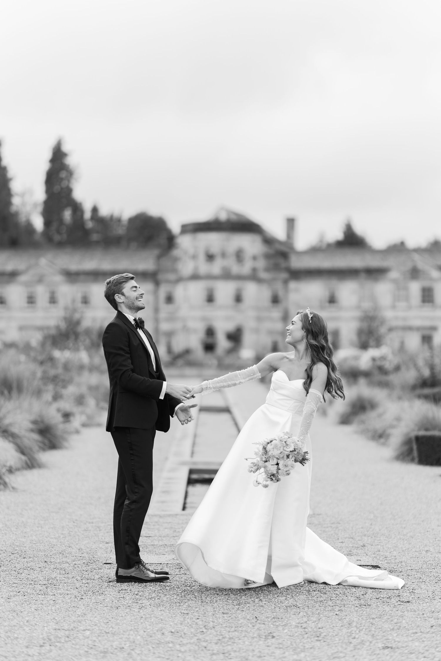 Bride and groom dancing joyfully on a gravel path in black and white outside a stately English manor.