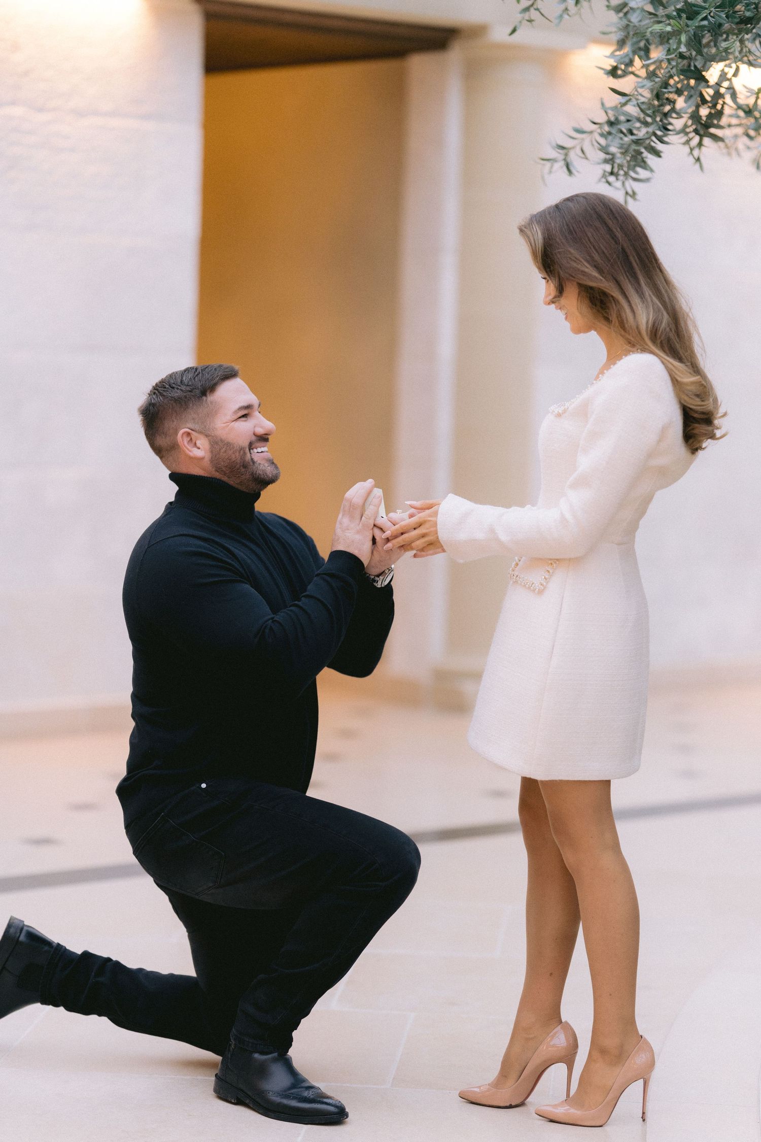 Man in black kneels proposing to woman in white mini dress, holding ring box outdoors.