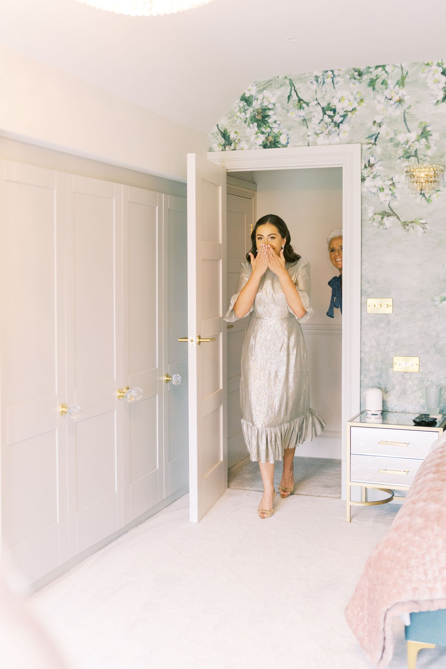 A surprised woman in a silver dress covers her mouth while standing in a doorway of an elegantly decorated room.