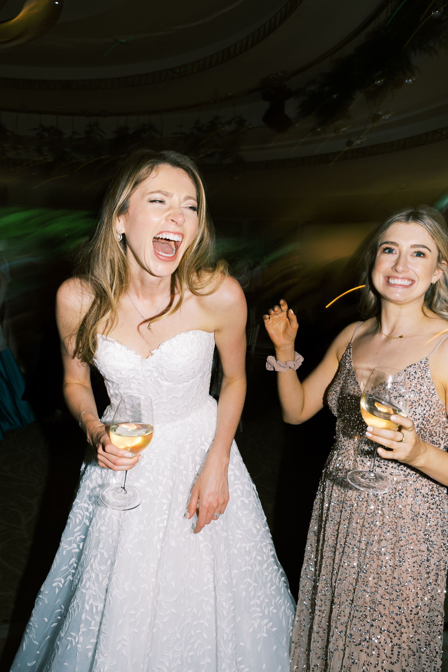 Joyful bride in white strapless gown laughing and dancing with bridesmaid in silver dress at wedding reception.