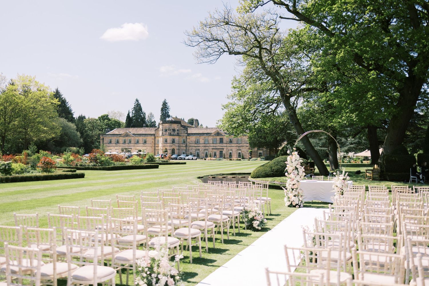 Elegant outdoor wedding ceremony setup with white chairs on a lawn, floral arch, and a grand manor house surrounded by trees.