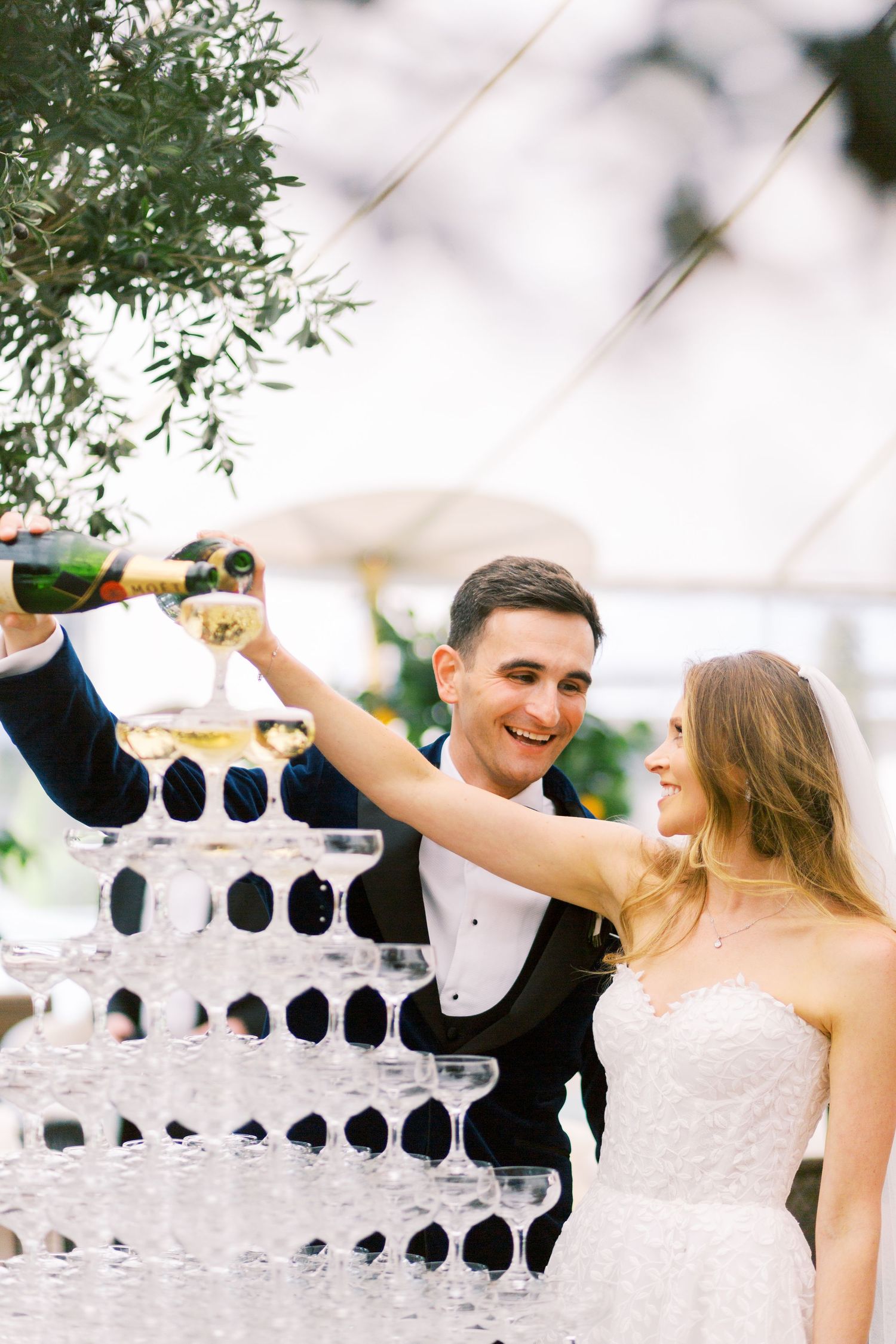 Smiling newlyweds pouring champagne into a elegant tiered champagne tower at their wedding reception.