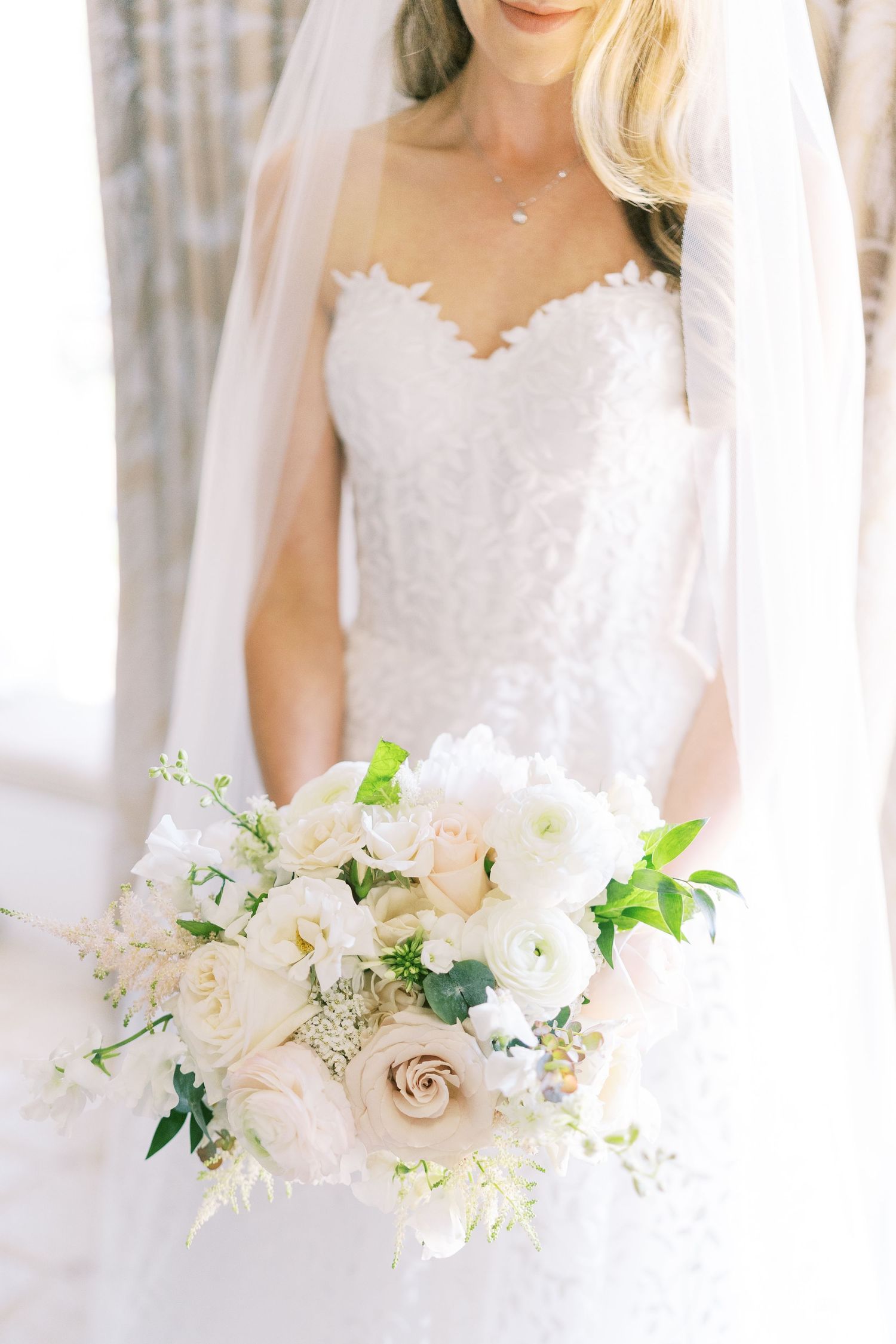 Bride in lace strapless gown holding white and blush floral bouquet with veil.