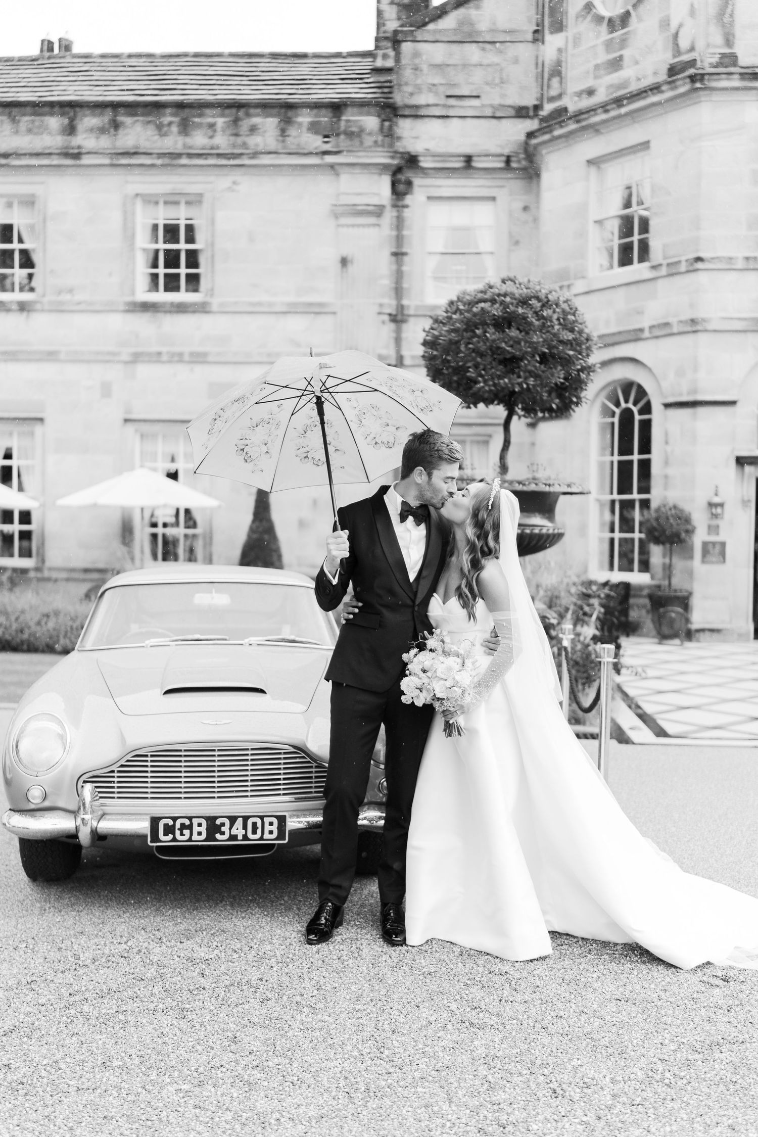 Bride and groom kissing beside a vintage Aston Martin with a lace umbrella outside an elegant stone manor.