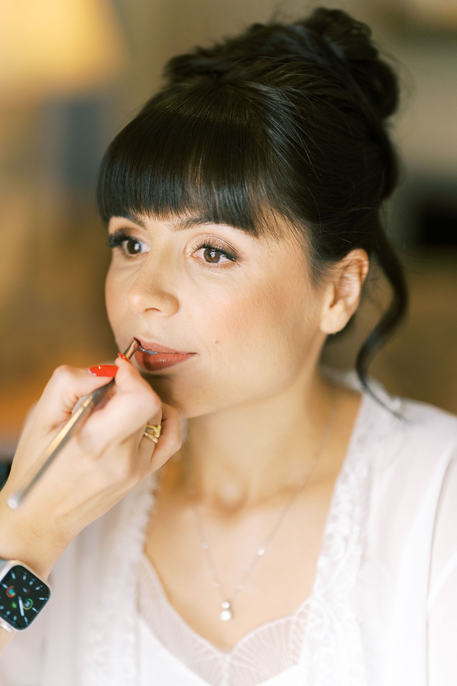 Makeup artist applying lipstick to dark-haired bride with updo hairstyle during wedding preparation.