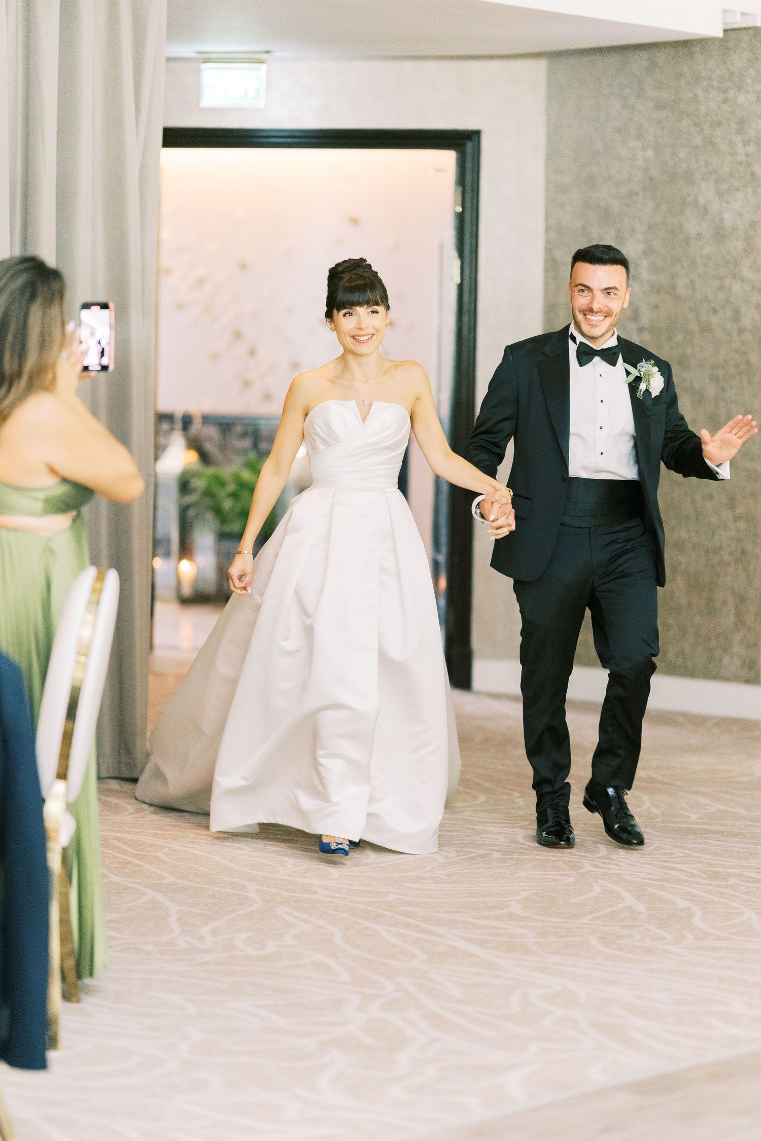 Smiling bride in strapless ball gown and groom in black tuxedo making grand entrance at wedding reception.