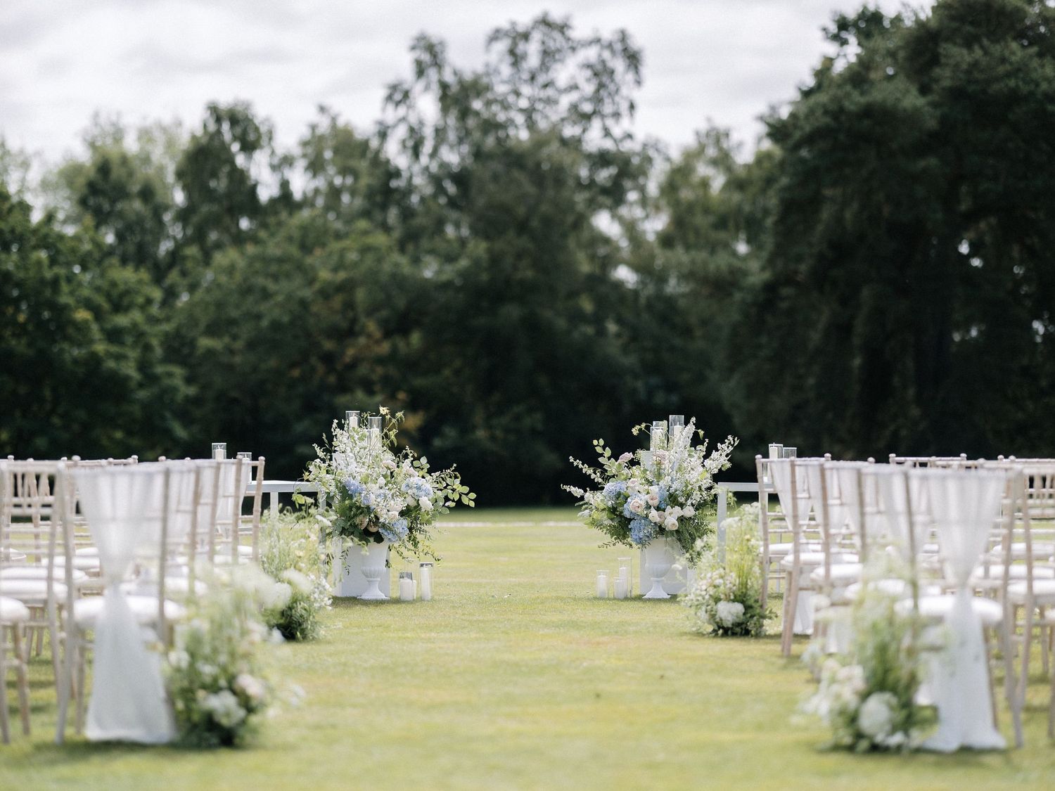 Elegant outdoor wedding aisle with white floral arrangements and chiavari chairs on green lawn.