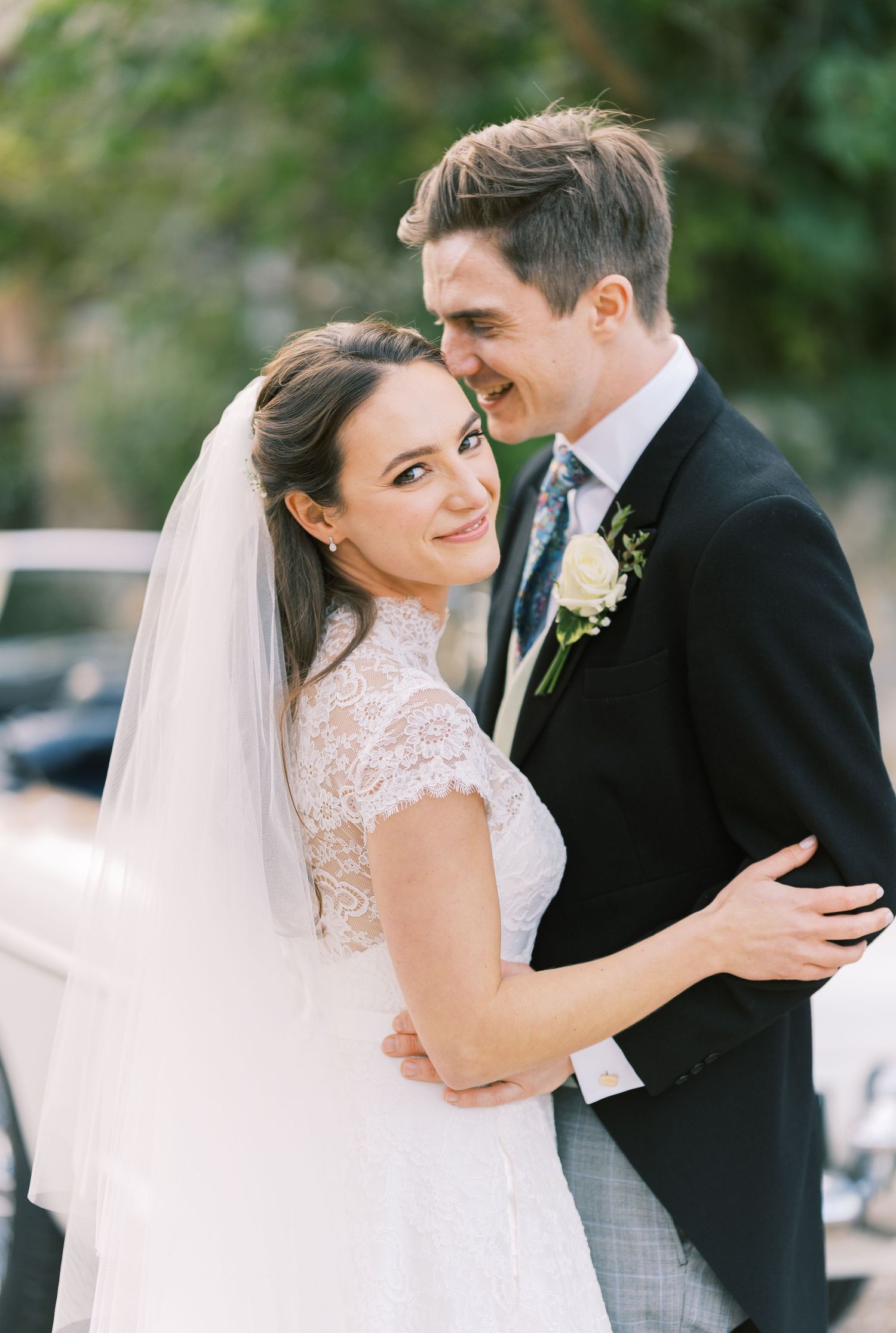 Smiling bride in lace gown with veil embracing groom in black suit outdoors.