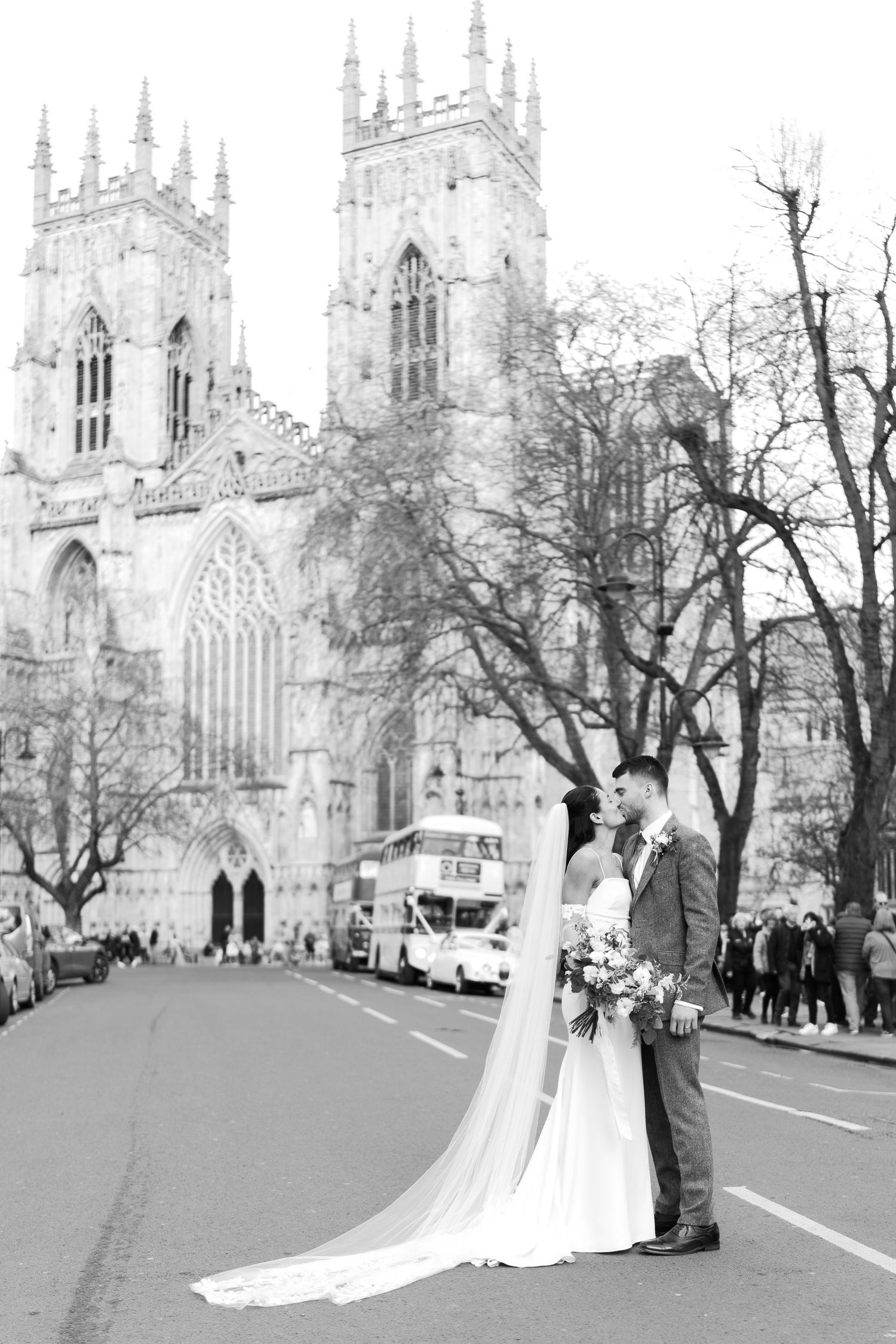 Black and white wedding photo of bride and groom kissing in front of York Minster Cathedral with a double-decker bus.