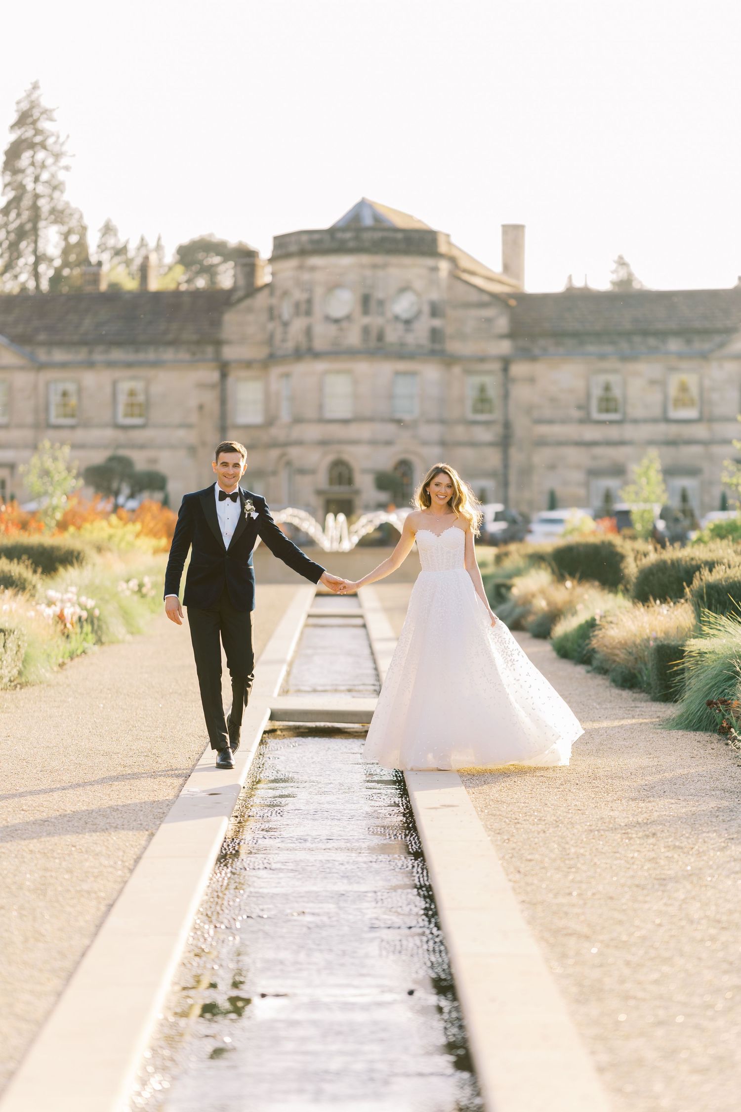 Bride and groom hold hands walking along a water feature with a grand estate venue in the background on their wedding day.