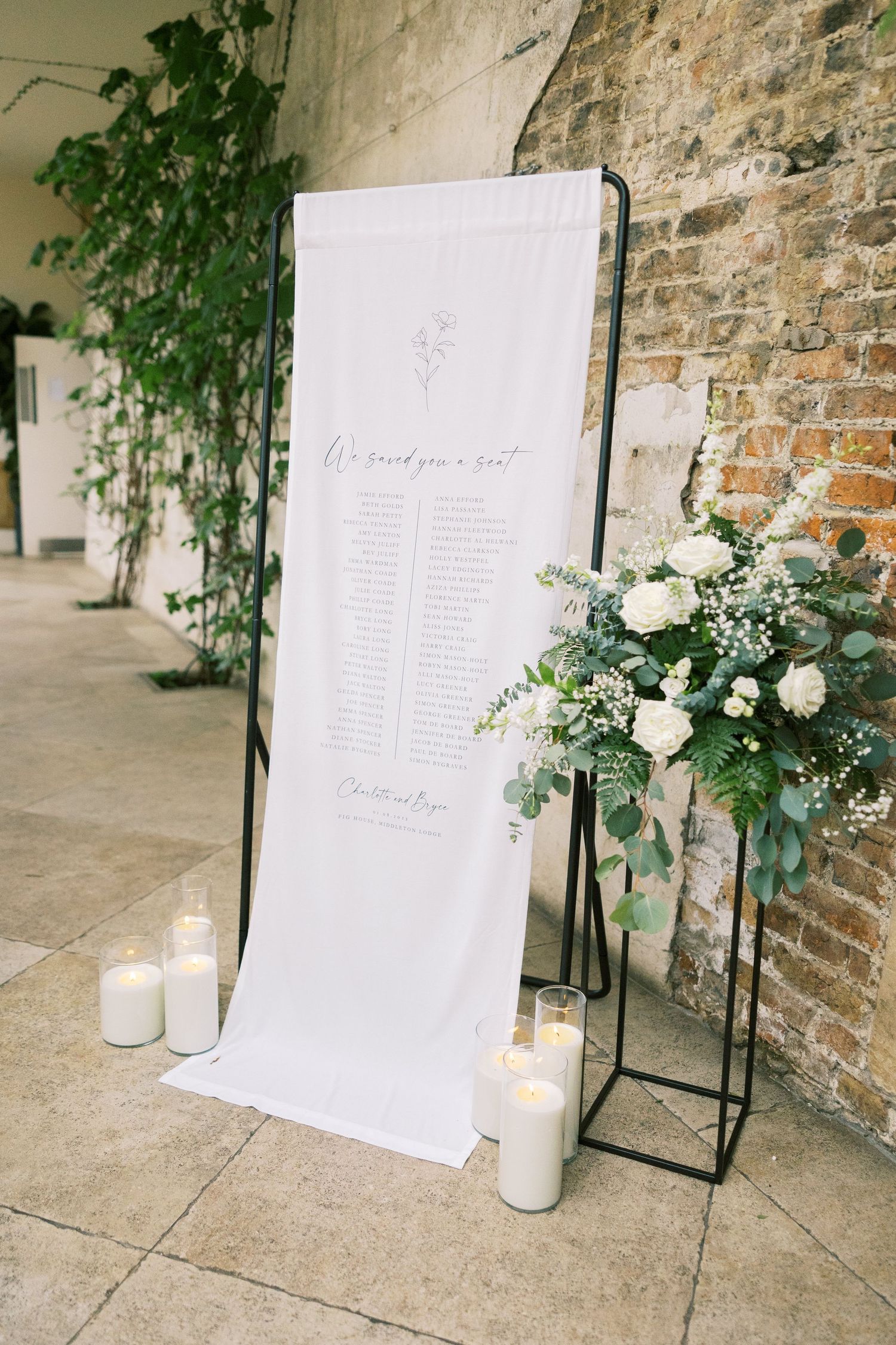 Elegant wedding seating chart on white fabric display with white floral arrangement and candles against rustic brick wall.