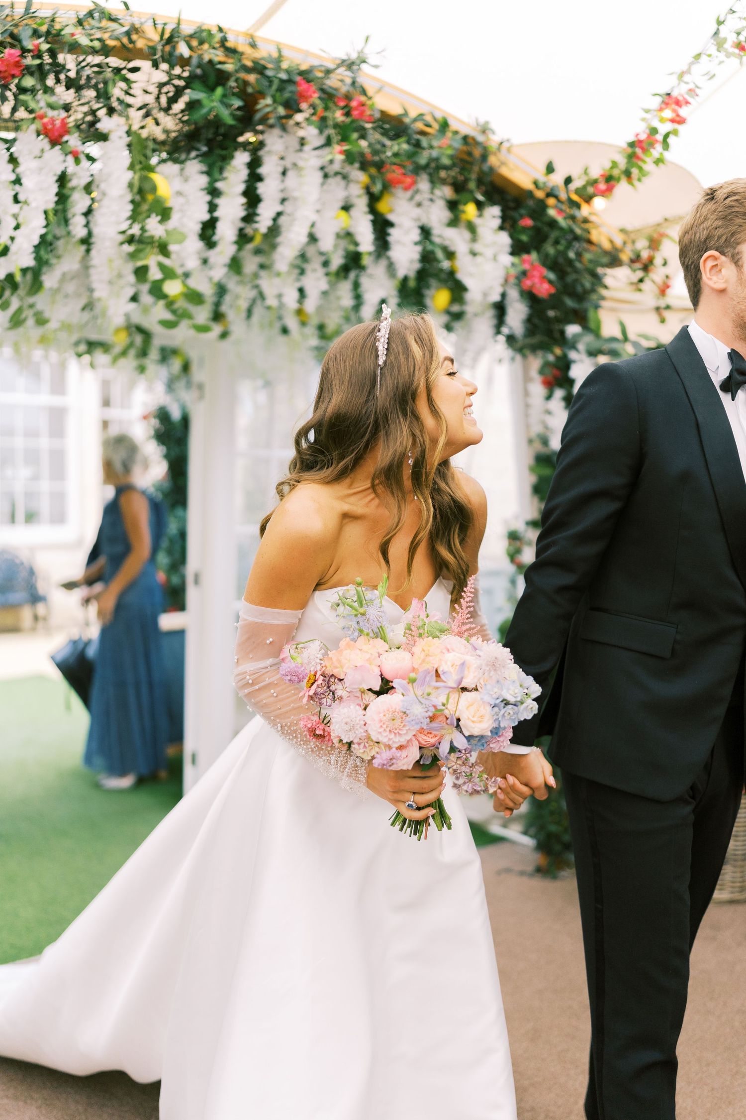 Bride in off-shoulder gown holds pastel bouquet beside groom in black tuxedo under floral arch at outdoor wedding.