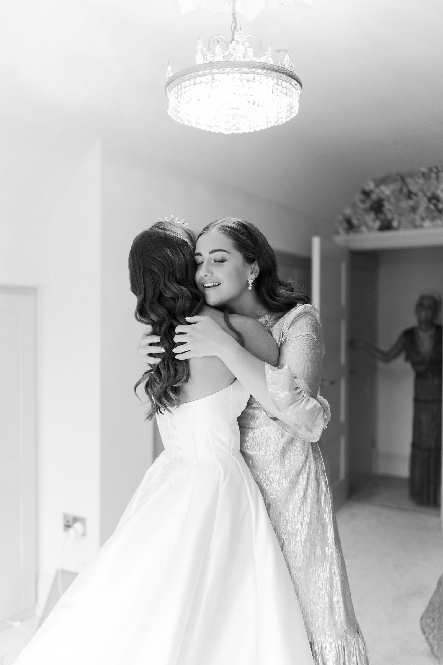 Bride in a white gown shares a heartfelt hug with a woman under a crystal chandelier during wedding preparations.