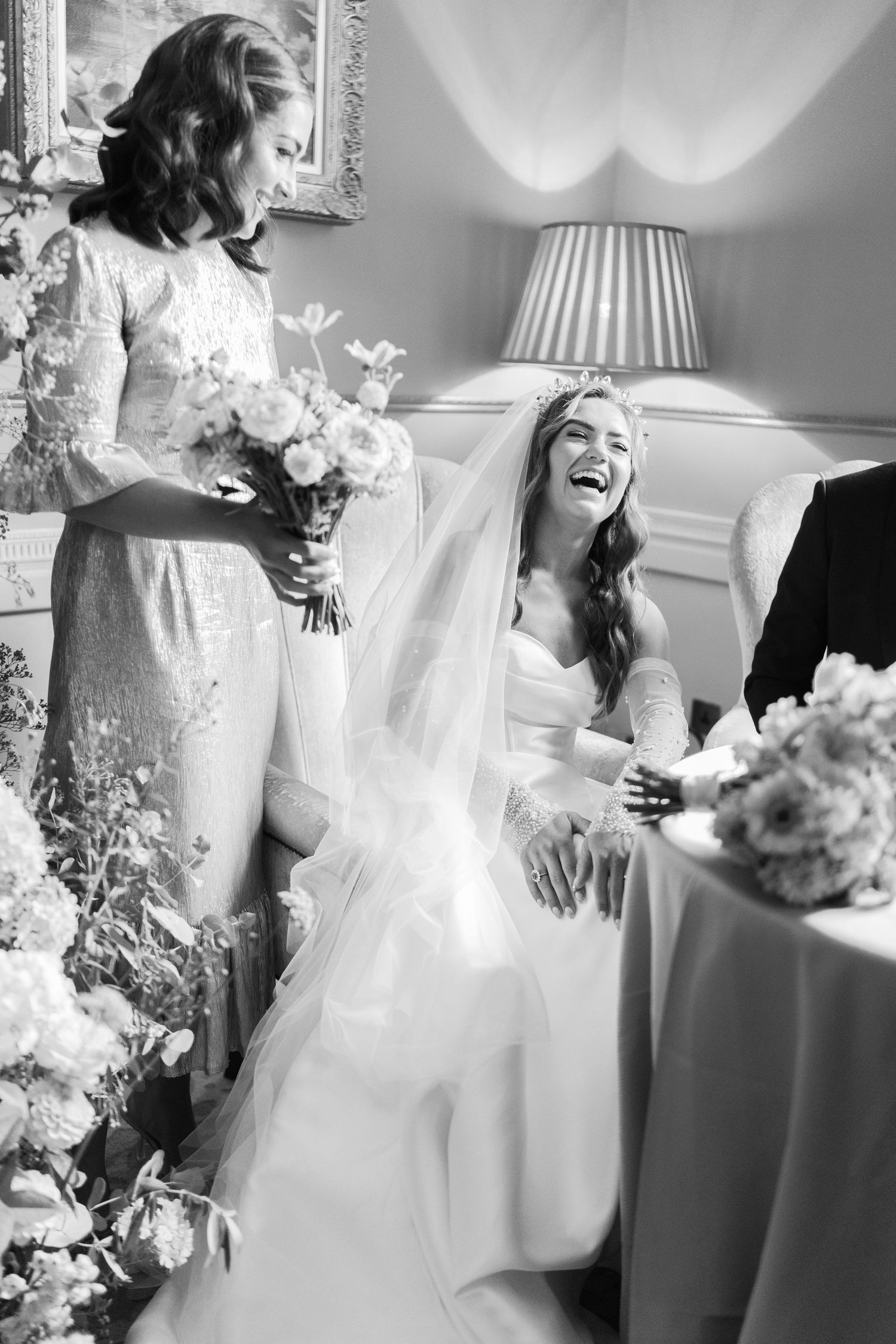 Bride laughing joyfully while seated in wedding dress and veil, bridesmaid holding bouquet beside her in elegant room.