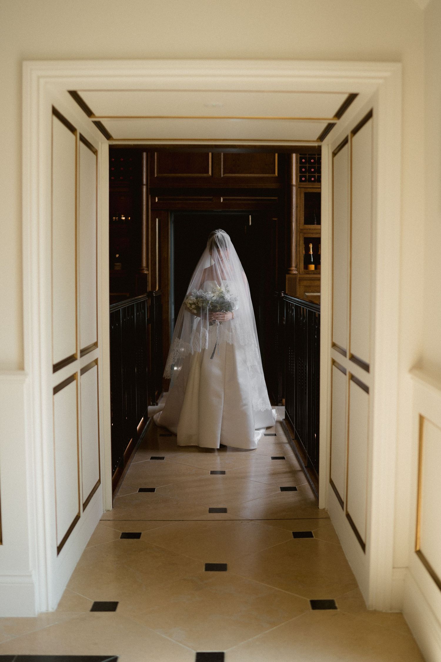 Bride in white gown and veil standing in elegant hallway doorway holding floral bouquet.