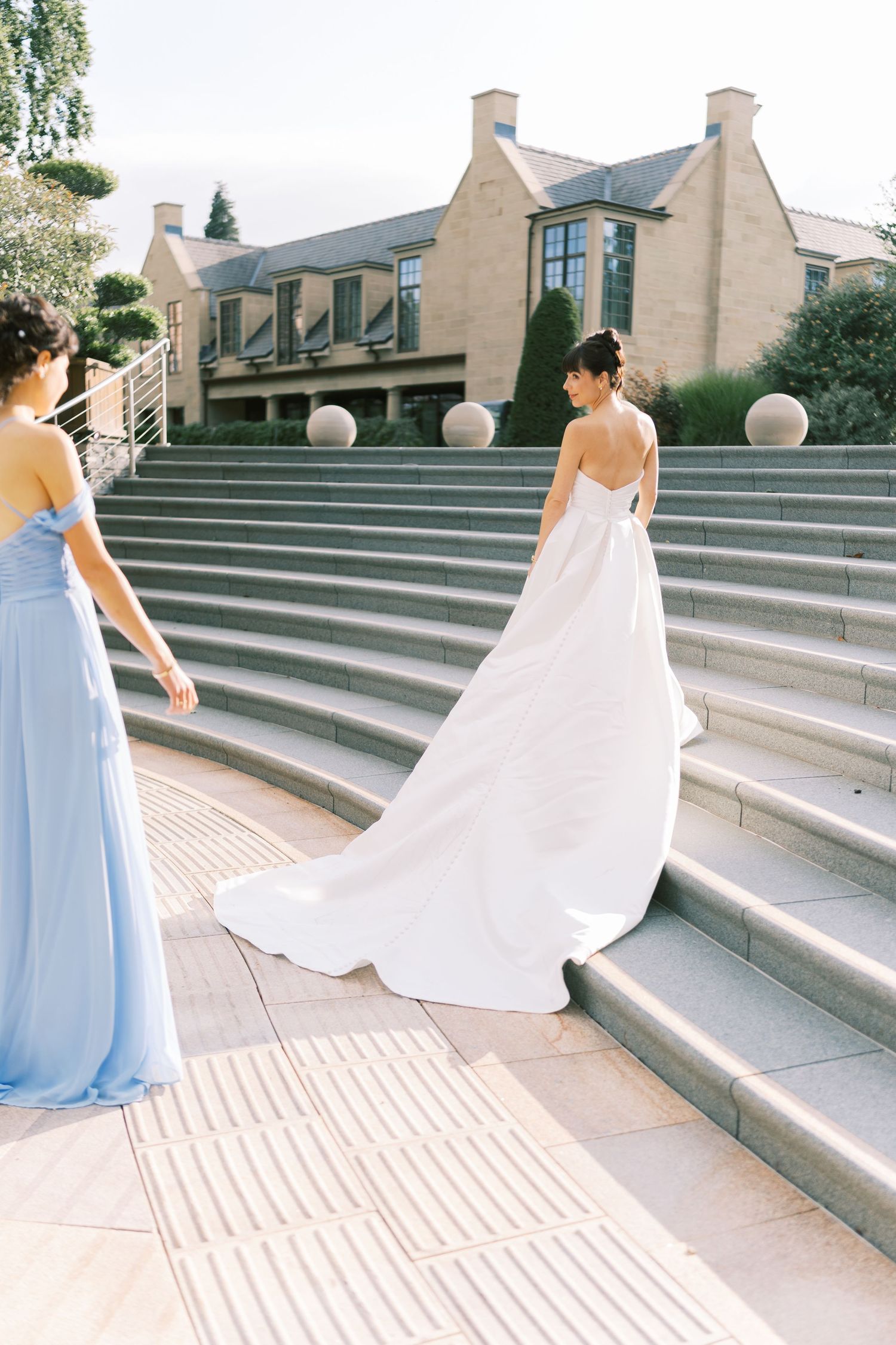Bride in white gown ascending stone steps with bridesmaid in blue dress at elegant venue.