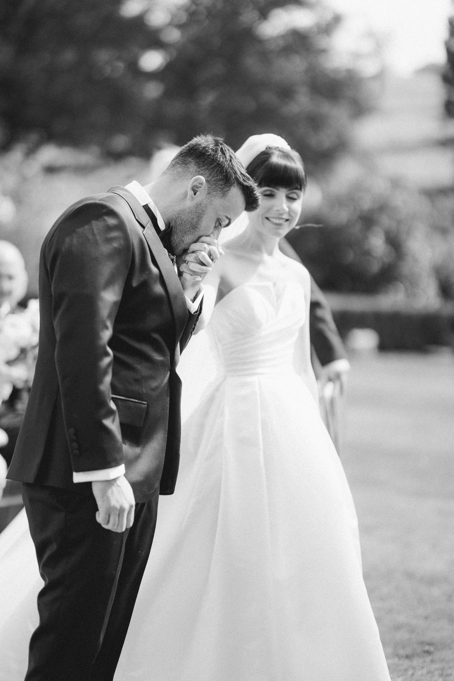 Groom kissing bride's hand during outdoor wedding ceremony in black and white photography.