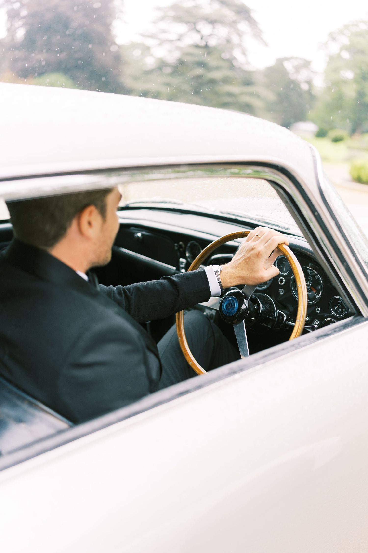 A groom in a dark suit drives a vintage classic car with a wooden steering wheel, wearing a watch on his wrist.