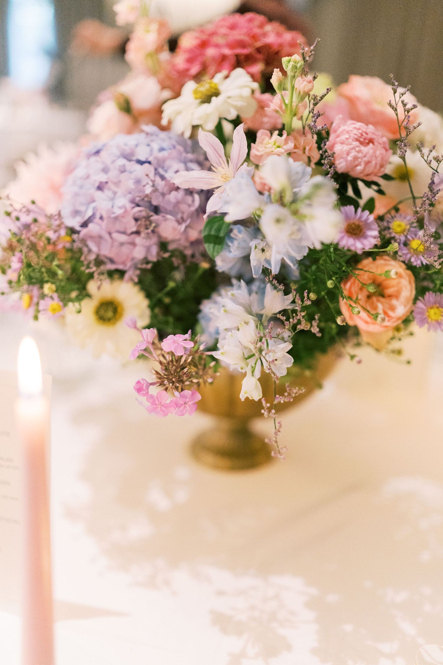 Elegant floral centerpiece with pink, purple hydrangeas, and white blooms in a gold vase beside a pink candle.