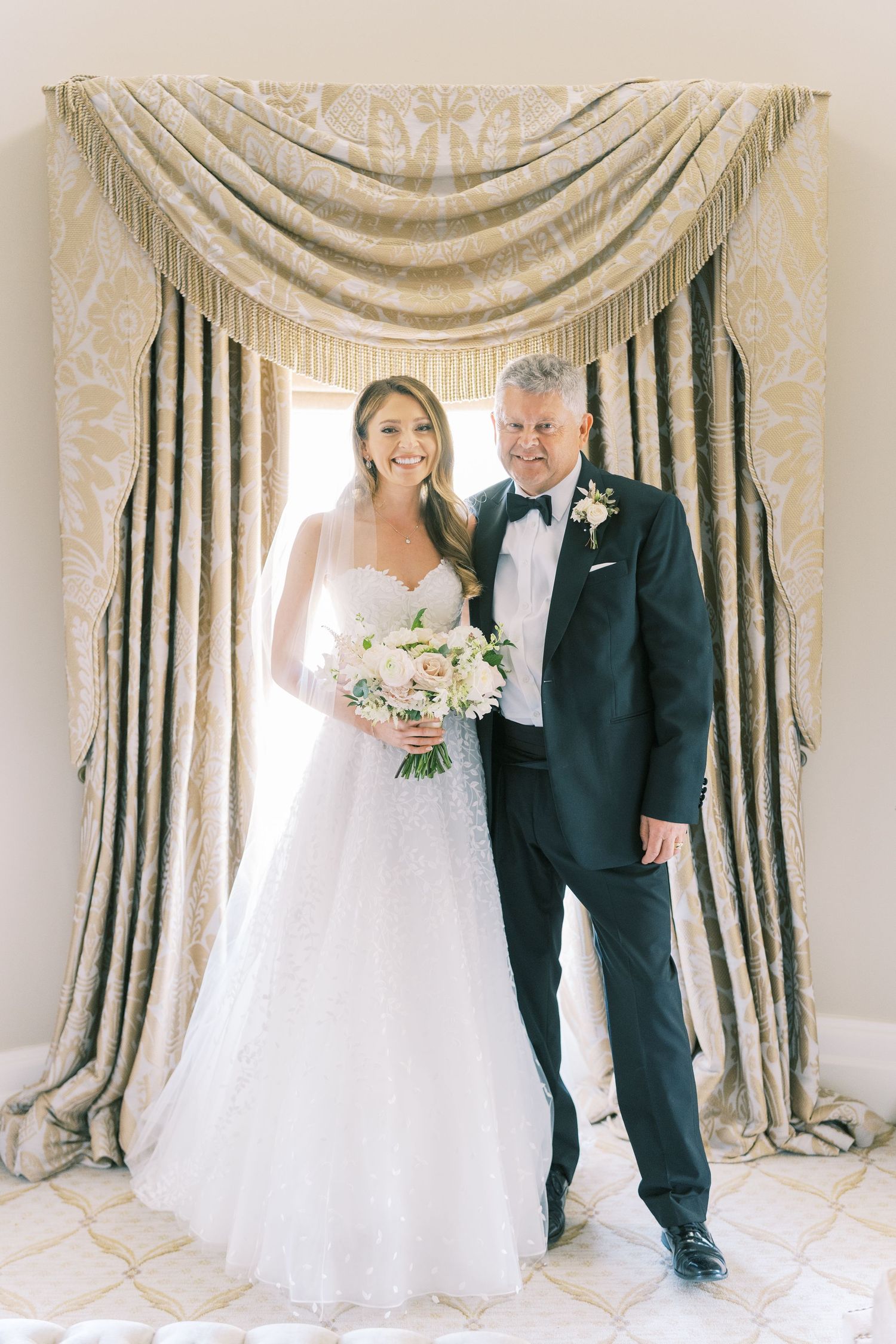 Bride in white gown holding bouquet poses with older man in tuxedo before elegant draped curtains.