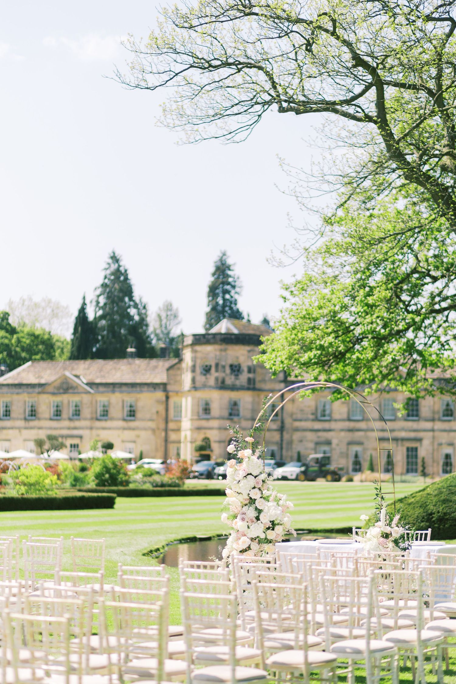 Elegant outdoor wedding ceremony setup with white chairs and floral arch on manicured lawn before a grand English manor.