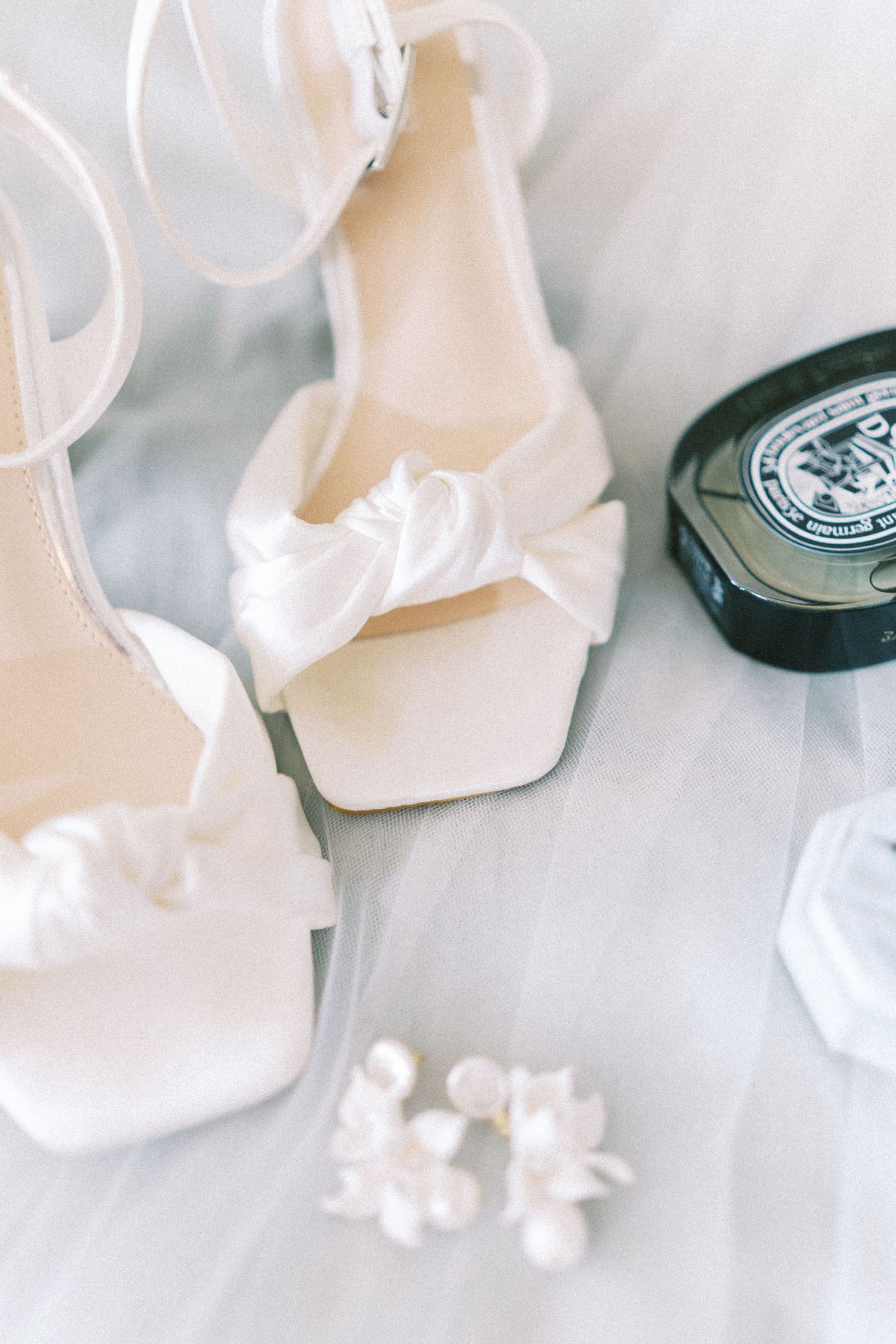 Elegant white satin bridal heels with bow detail beside pearl earrings and perfume on soft fabric.