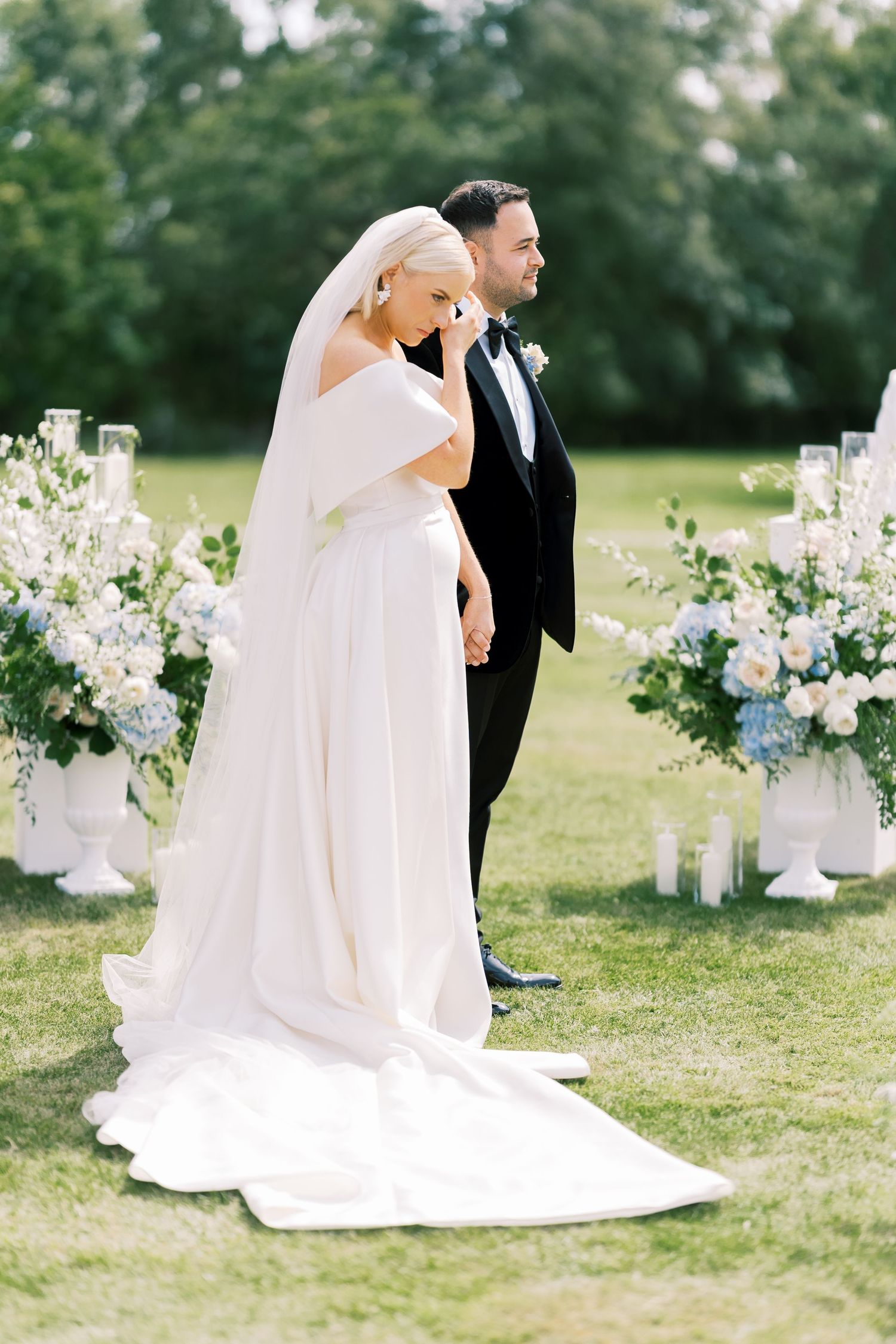 Bride in off-shoulder white gown with veil standing beside groom at outdoor wedding ceremony.