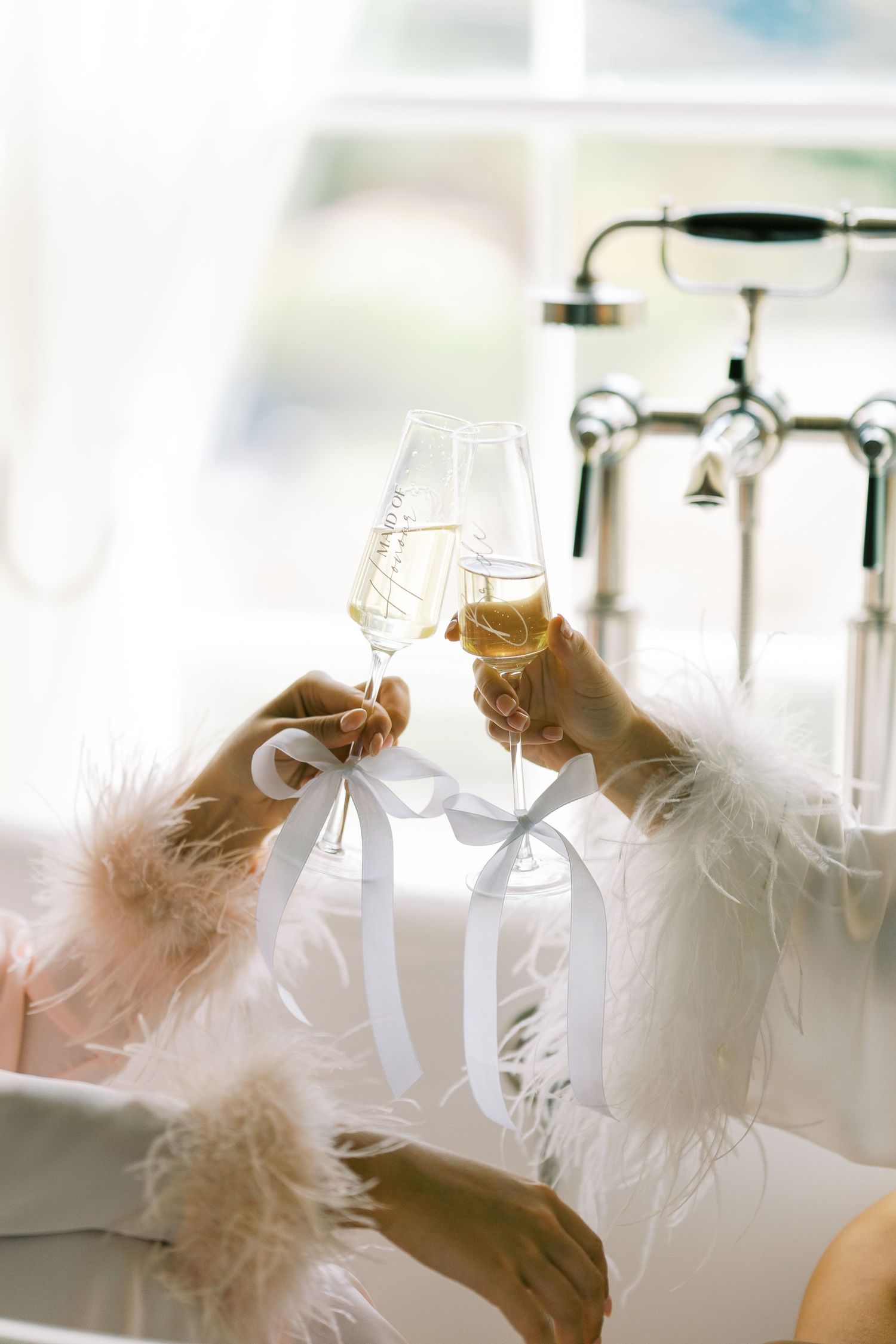 Two women in feather robes toasting with champagne flutes tied with white ribbons.
