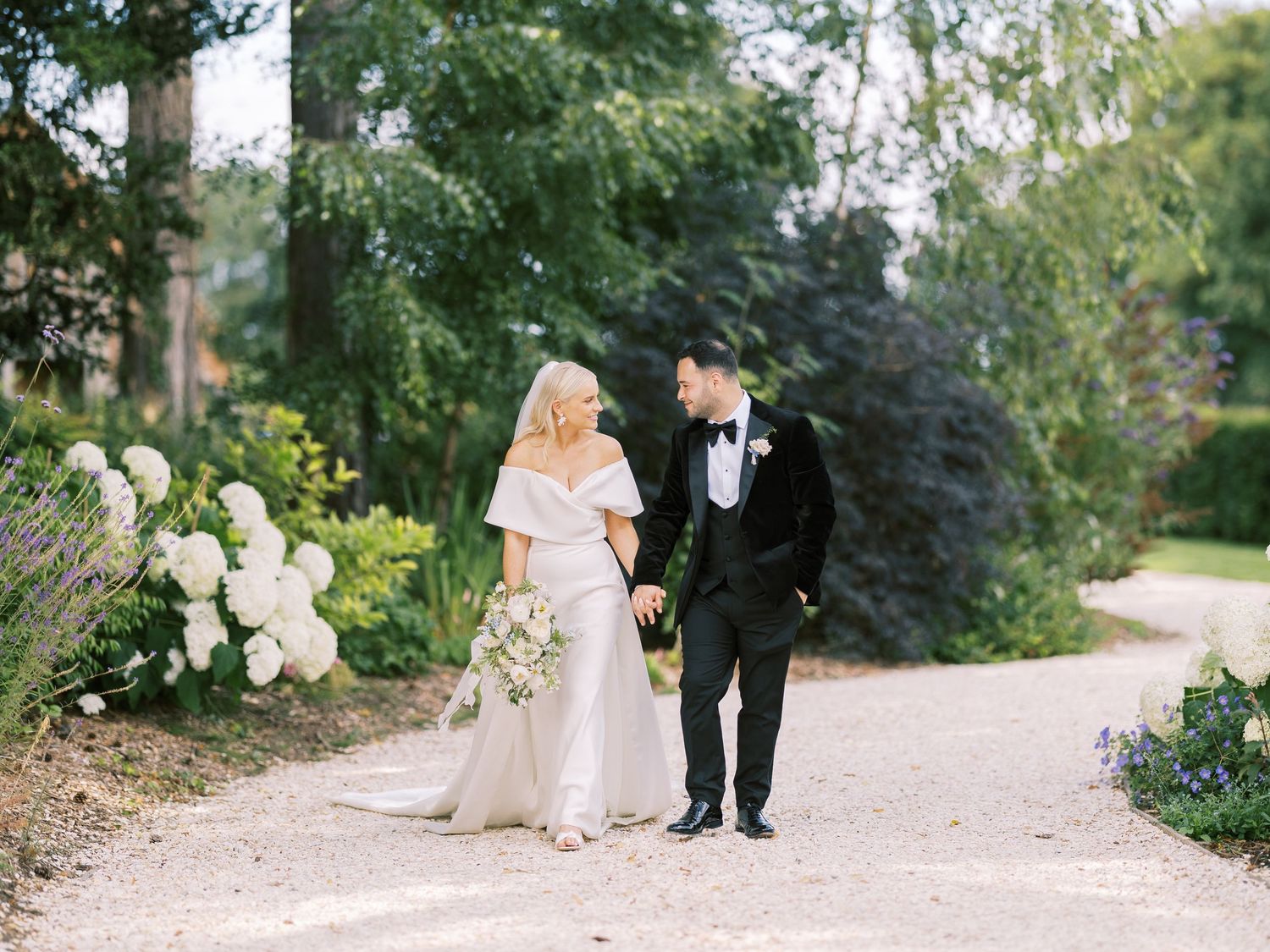 Bride and groom walk hand-in-hand along garden path lined with hydrangeas and lush greenery.