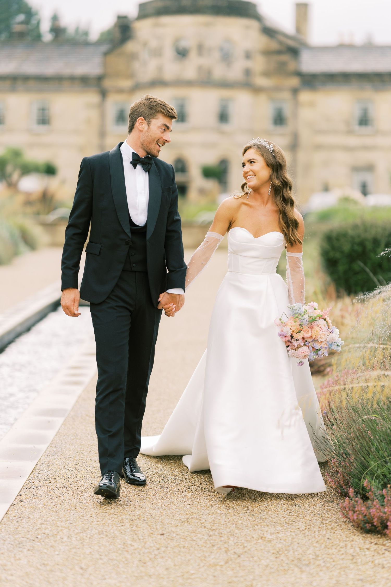 Smiling bride and groom walk hand-in-hand outside elegant stone manor, she carries a pastel bouquet.