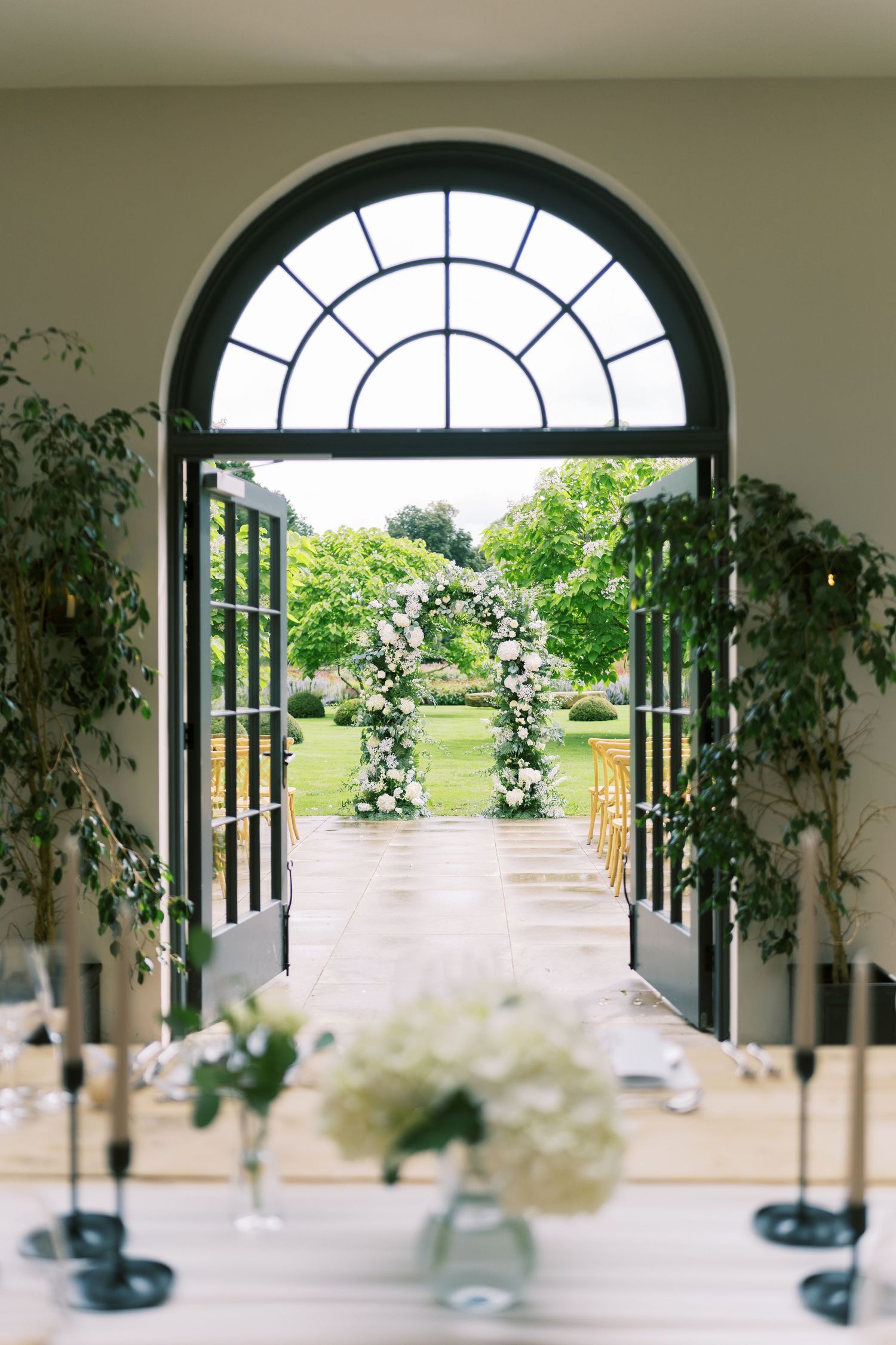 Elegant wedding venue with arched window doors opening to a floral arch ceremony setup in a lush green garden.