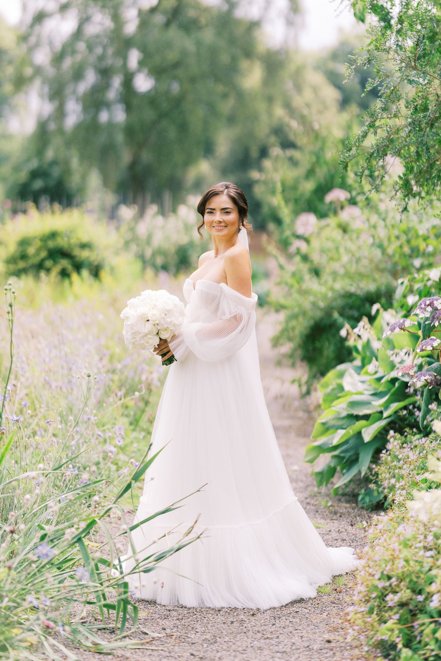 Smiling bride in off-shoulder white gown holds bouquet while walking through lush garden path.