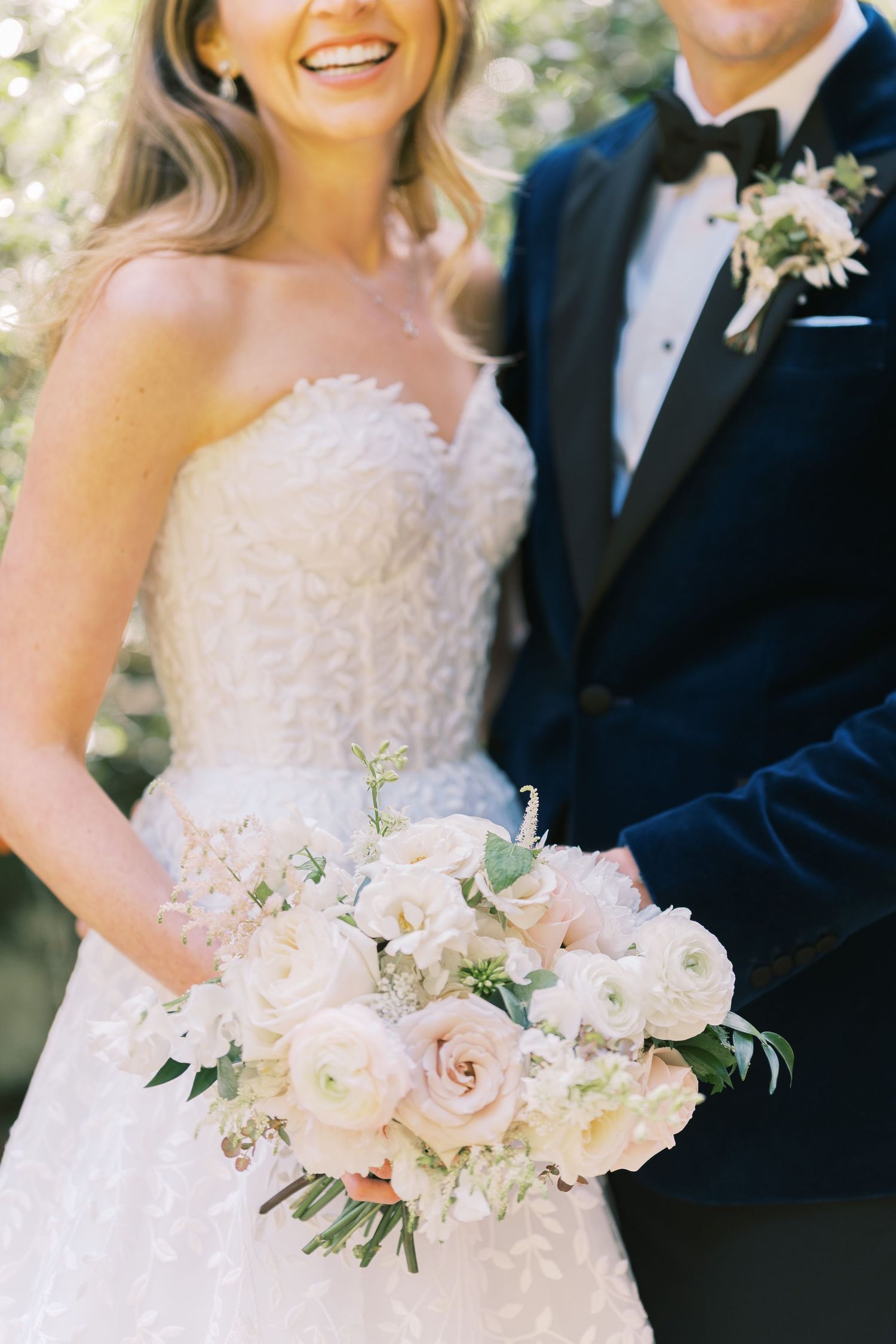 Smiling bride in strapless lace gown holds white and blush floral bouquet beside groom in navy tuxedo.