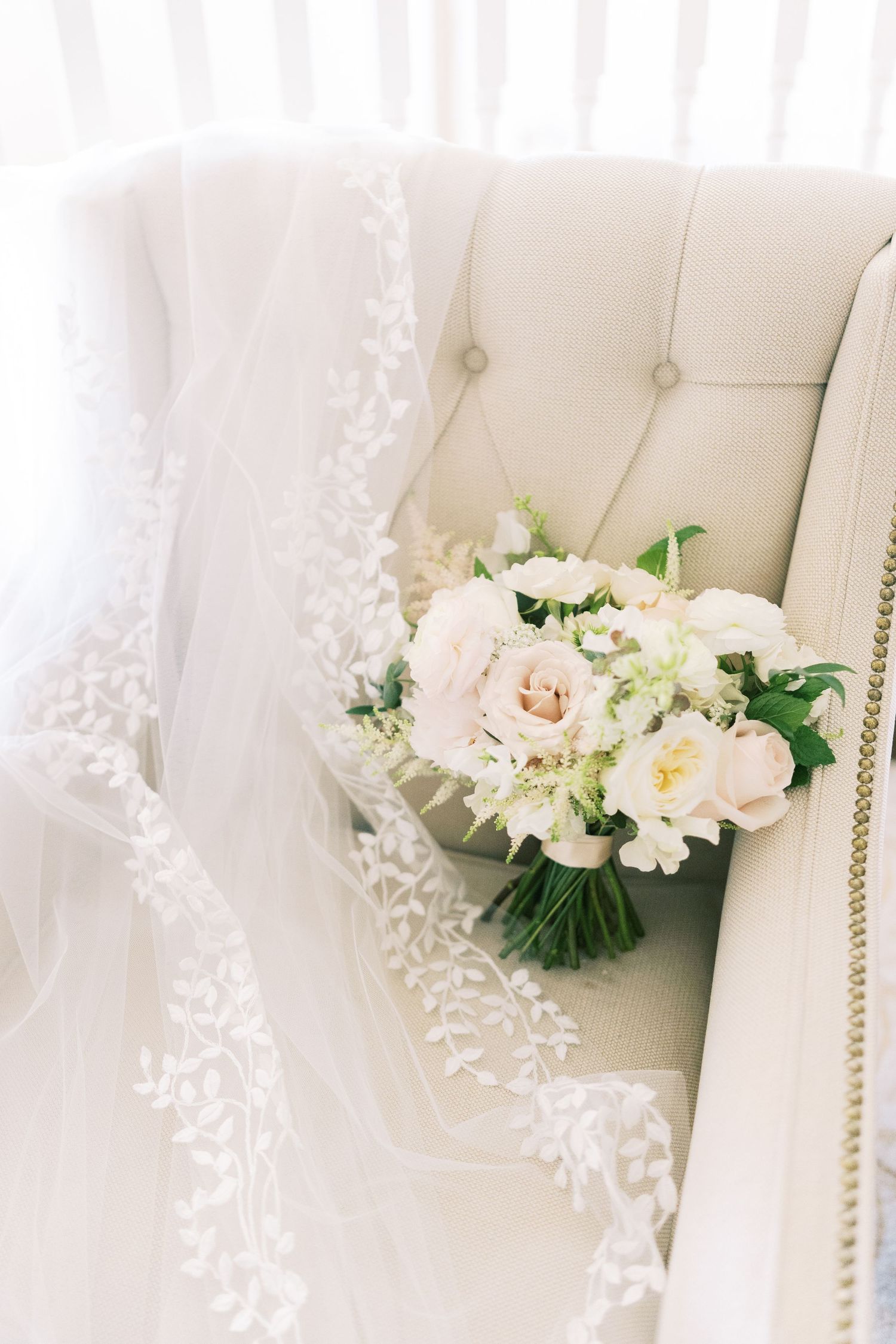 Bridal bouquet of blush roses and white flowers resting on a tufted ivory chair beside a lace wedding veil.