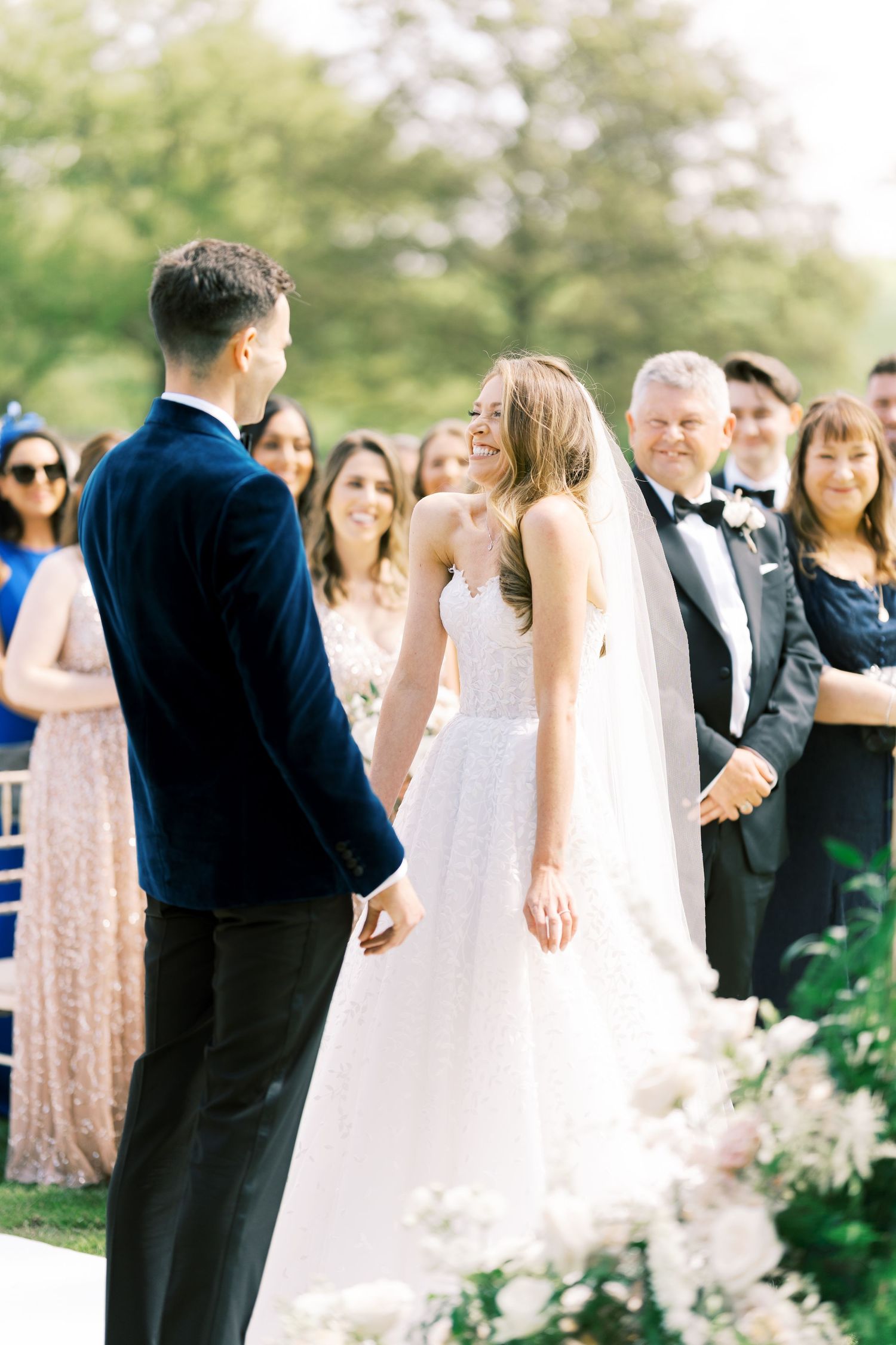 Bride smiling joyfully at groom during outdoor wedding ceremony, wearing white ballgown and veil, guests watching.