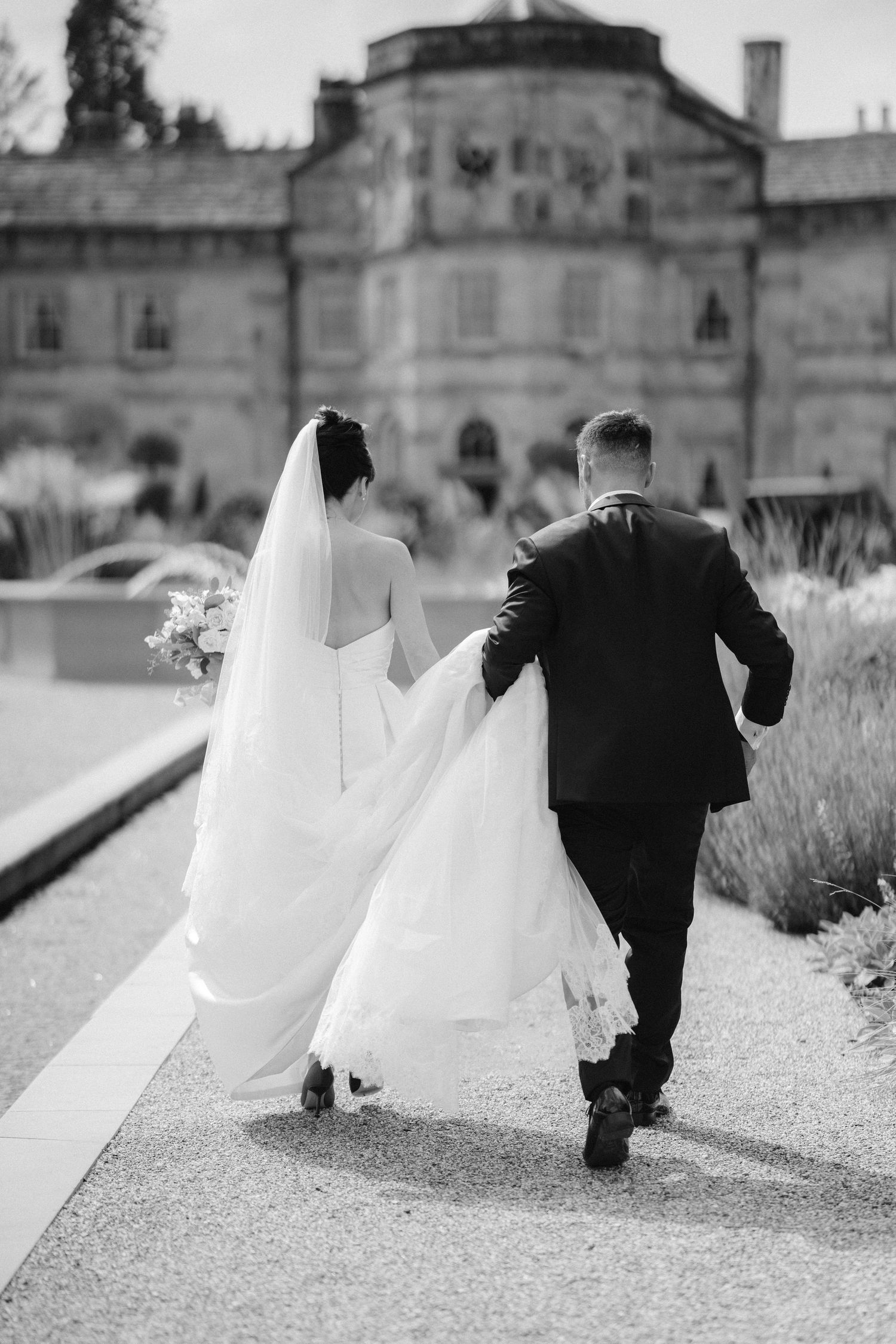 Black and white photo of bride and groom walking hand in hand toward elegant estate building on their wedding day.