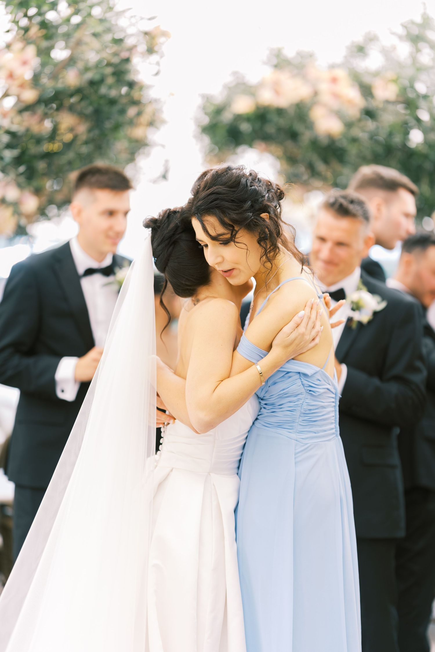 Bride and bridesmaid share an emotional hug during outdoor wedding ceremony with groomsmen in background.